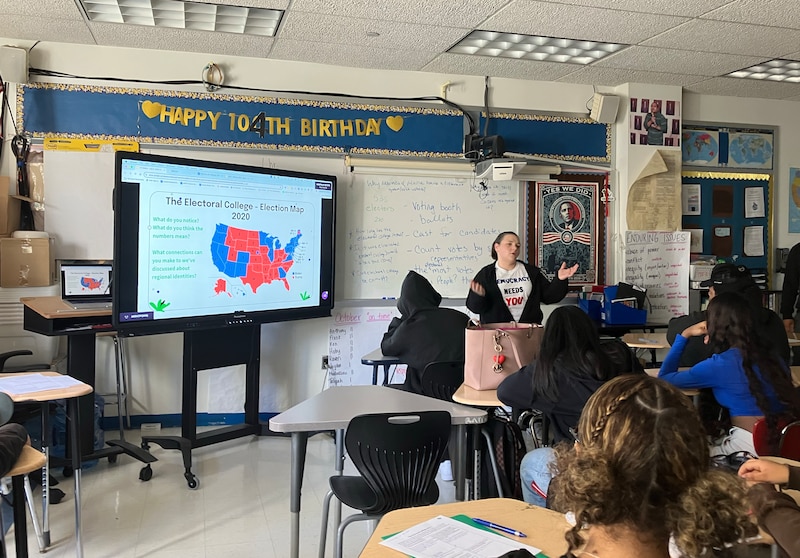 A teacher stands at the front of a class while students sit in rows of desks.