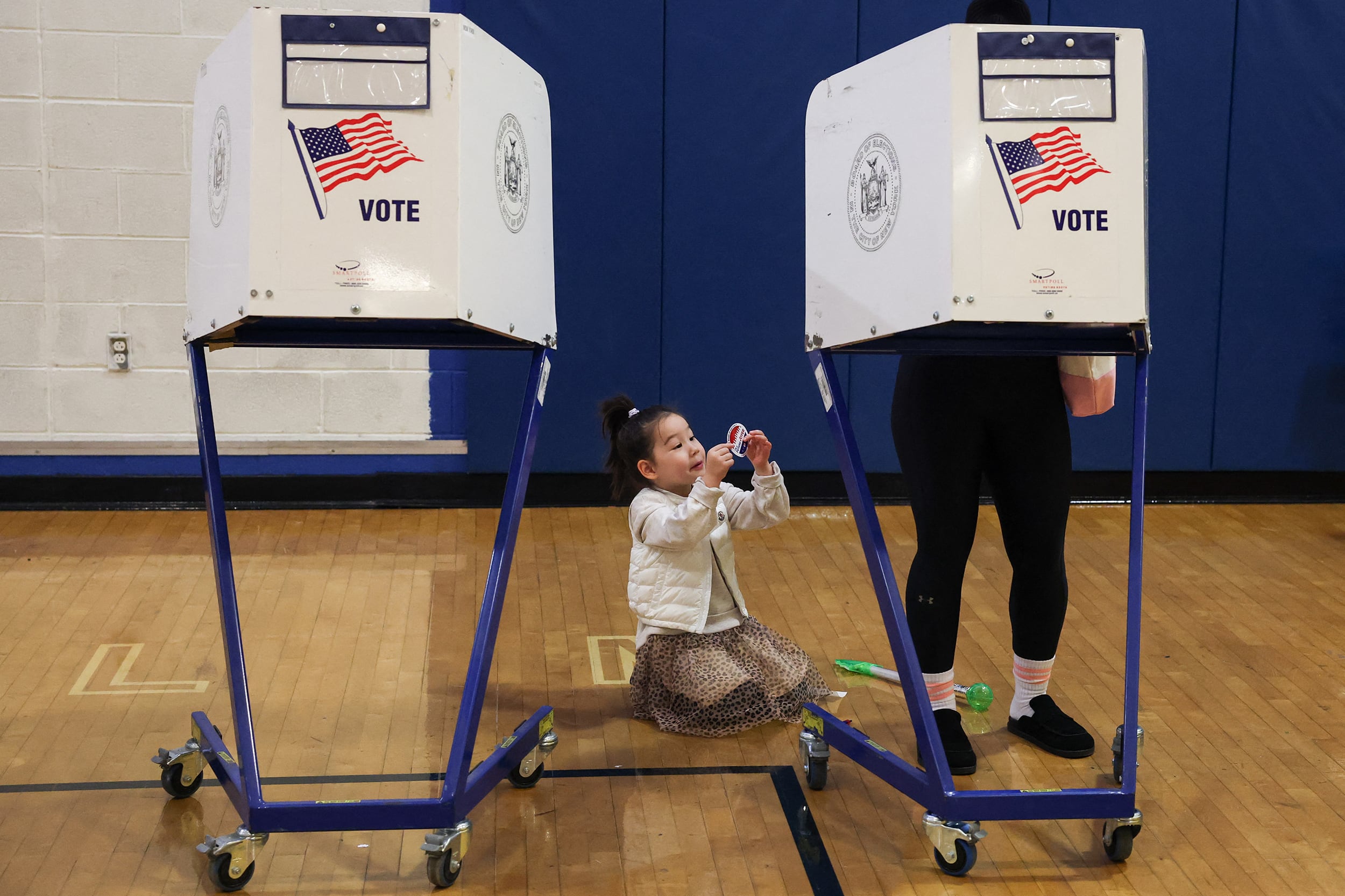 A photograph of a child sitting on the floor while a person stands behind a voting divider in a school gym.