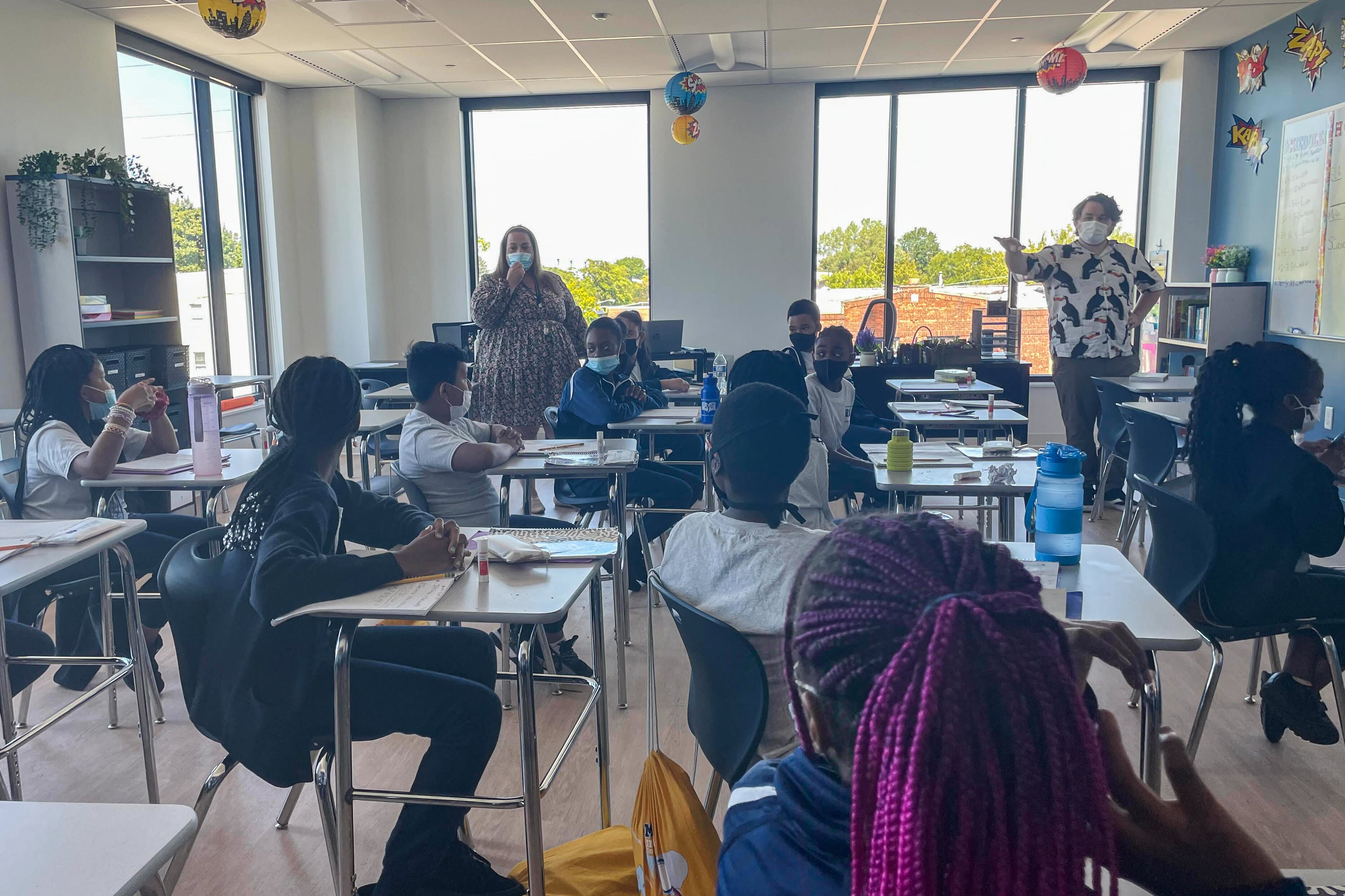 A classroom of students sit at their desks as a teacher in a dress asks them questions.