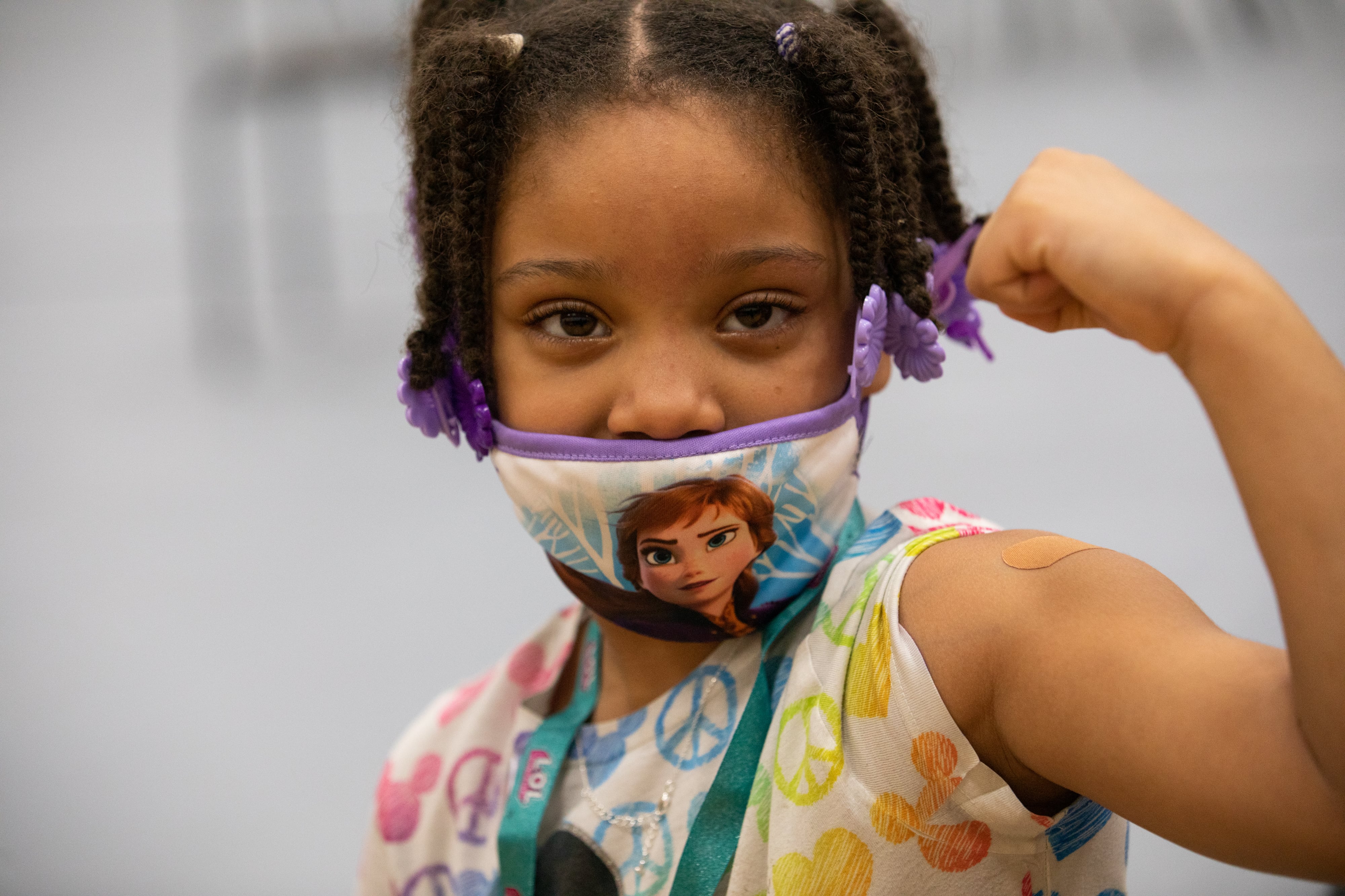 A young girl wearing a mask looks directly at the camera and holds up her arm to show off her bandage after getting a COVID-19 vaccine shot.