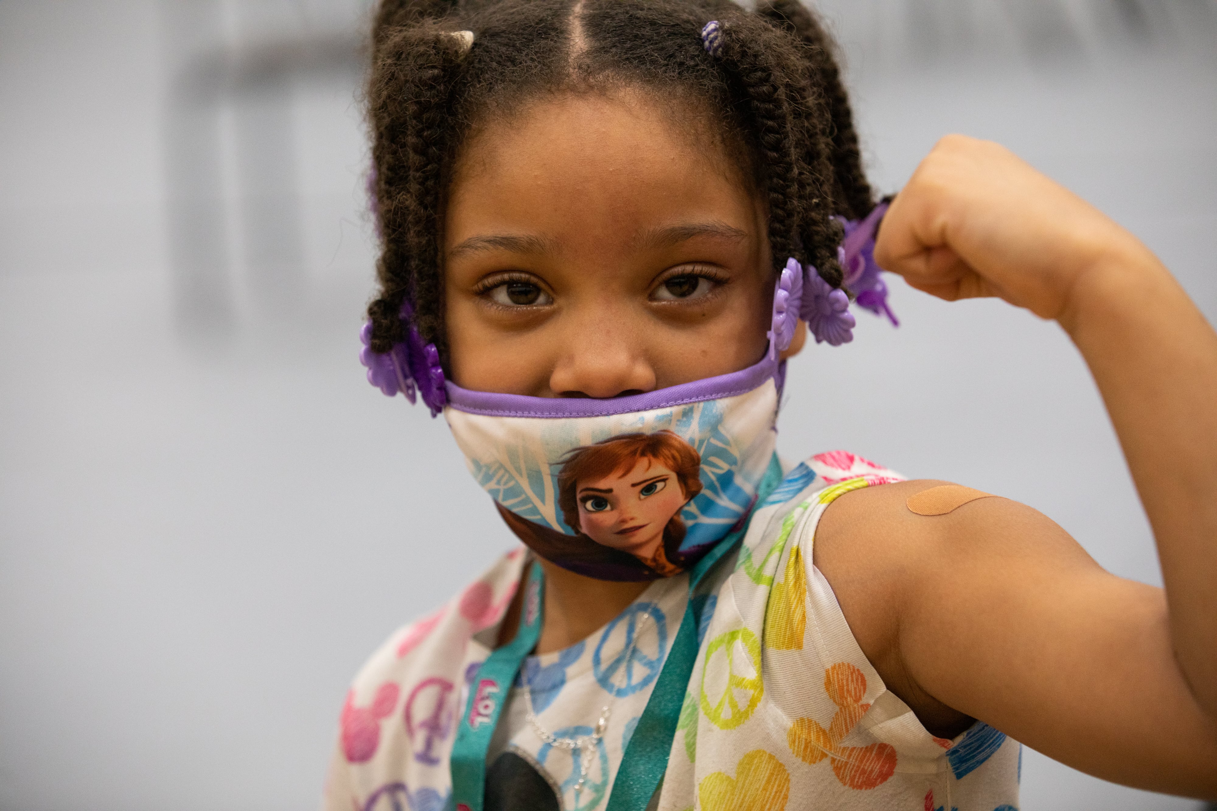 A young girl wearing a mask looks directly at the camera and holds up her arm to show off her bandage after getting a COVID-19 vaccine shot.