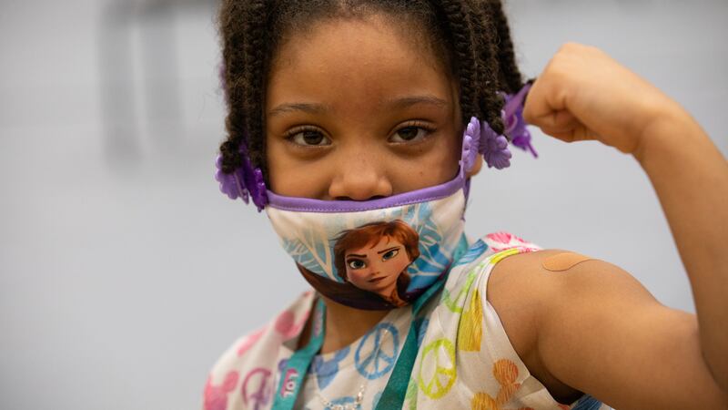 A young girl wearing a mask looks directly at the camera and holds up her arm to show off her bandage after getting a COVID-19 vaccine shot.