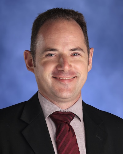 Portrait of Lem Wheeles. Wheeles is wearing a dark blazer and a red striped tie. He has short hair and he's smiling at the camera.