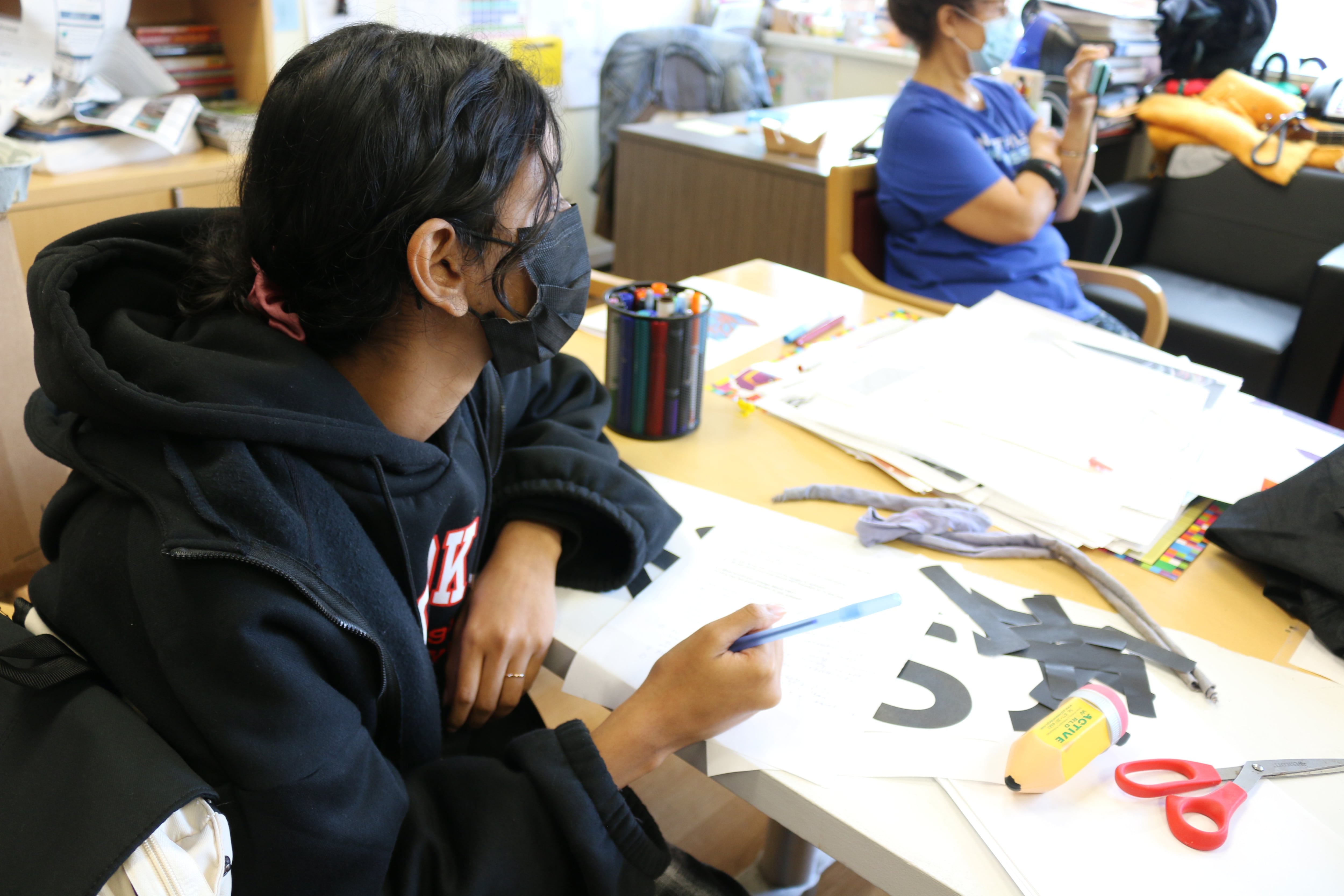 A student wearing a black hoodie and protective mask works at their desk during class.