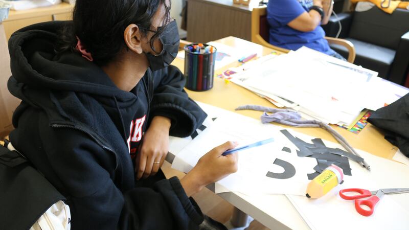 A student wearing a black hoodie and protective mask works at their desk during class.