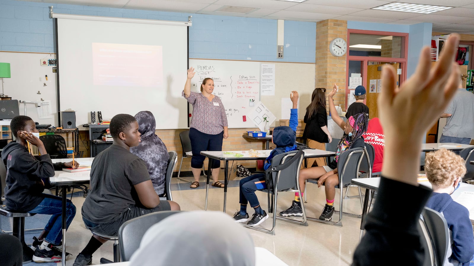 A woman teacher leads her classroom during a lesson, a student in the foreground raising their hand amongst other students in the class.