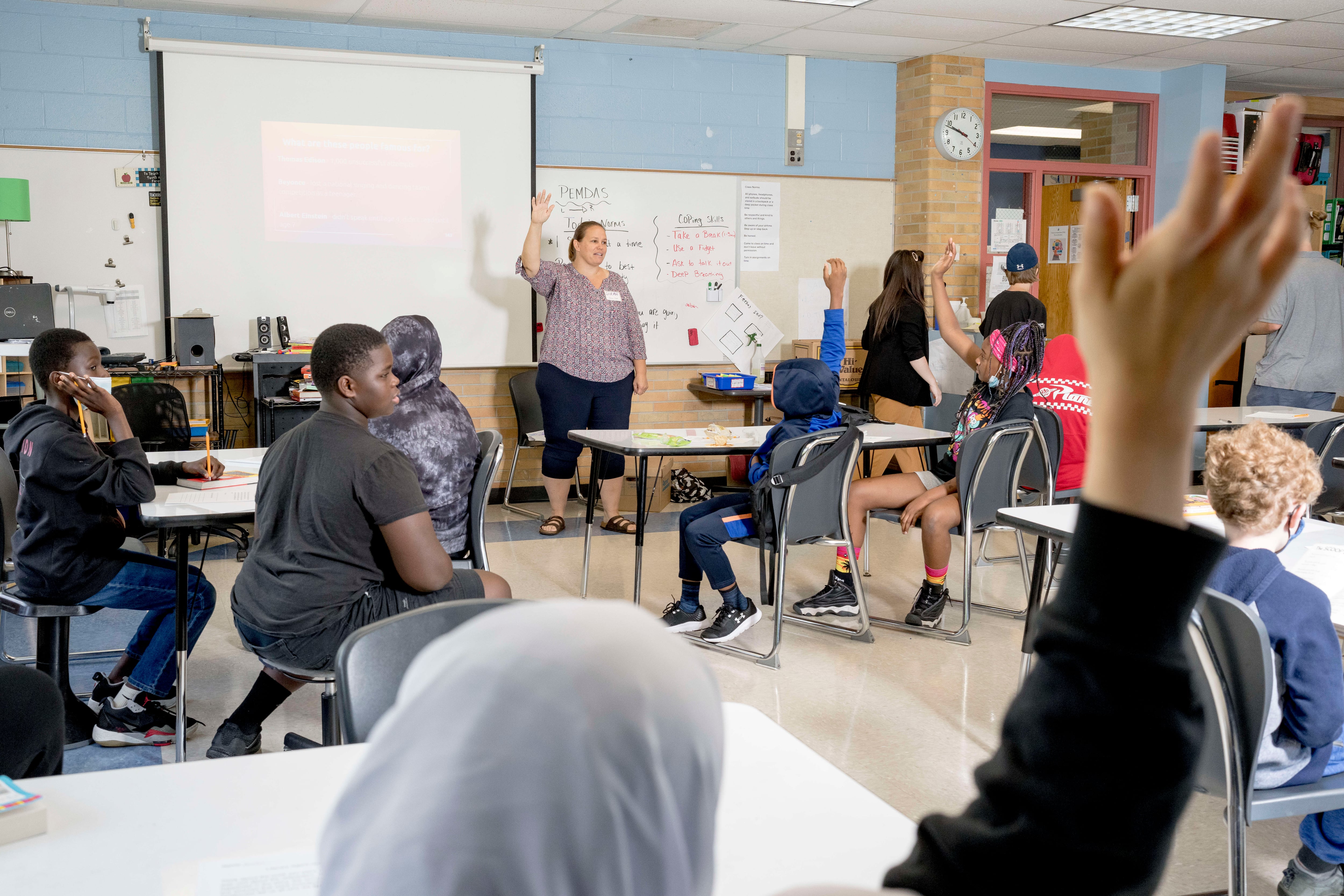A woman teacher leads her classroom during a lesson, a student in the foreground raising their hand amongst other students in the class.