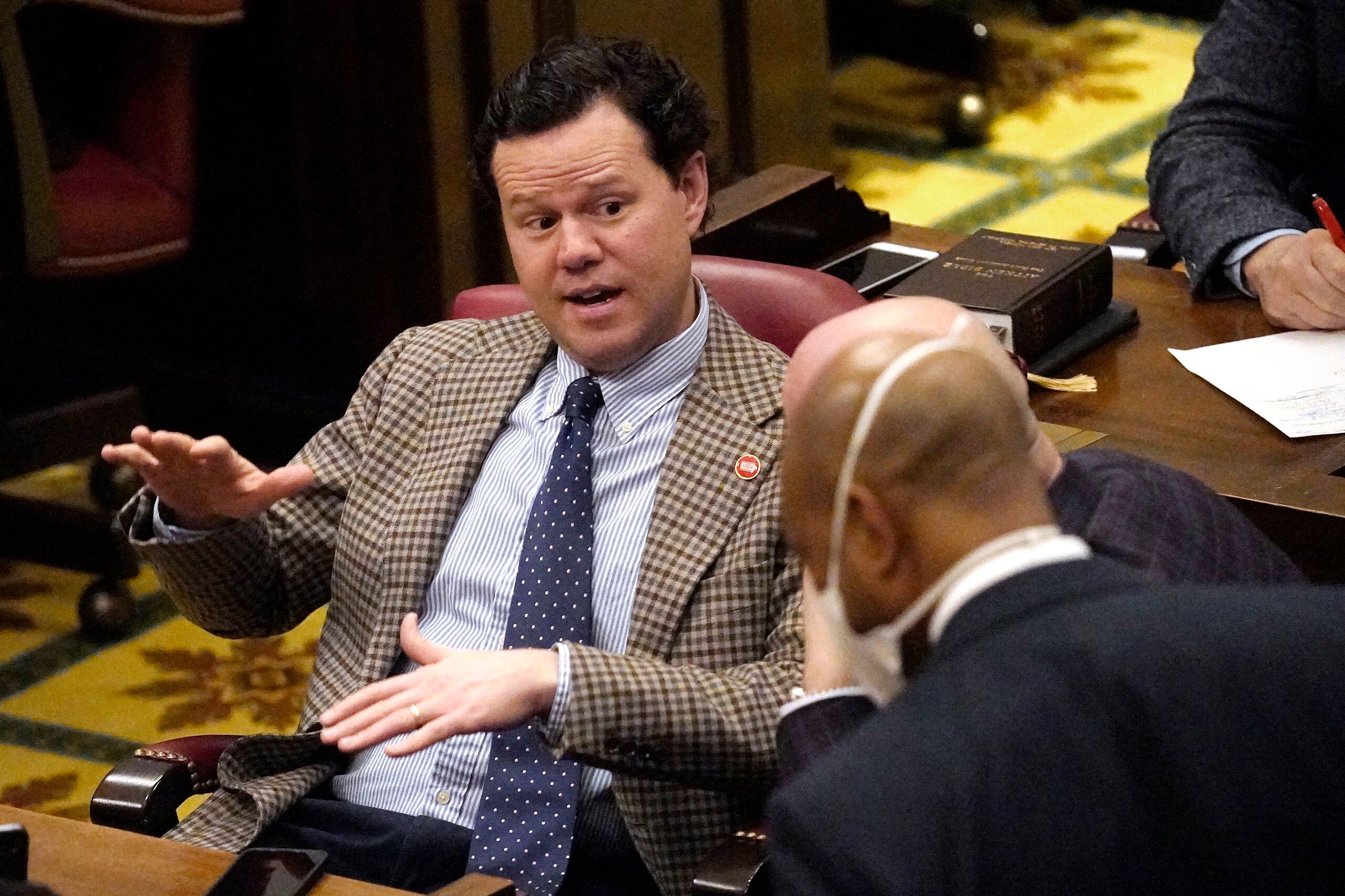 A man wearing a suit and tie speaks with two other men in an assembly room.