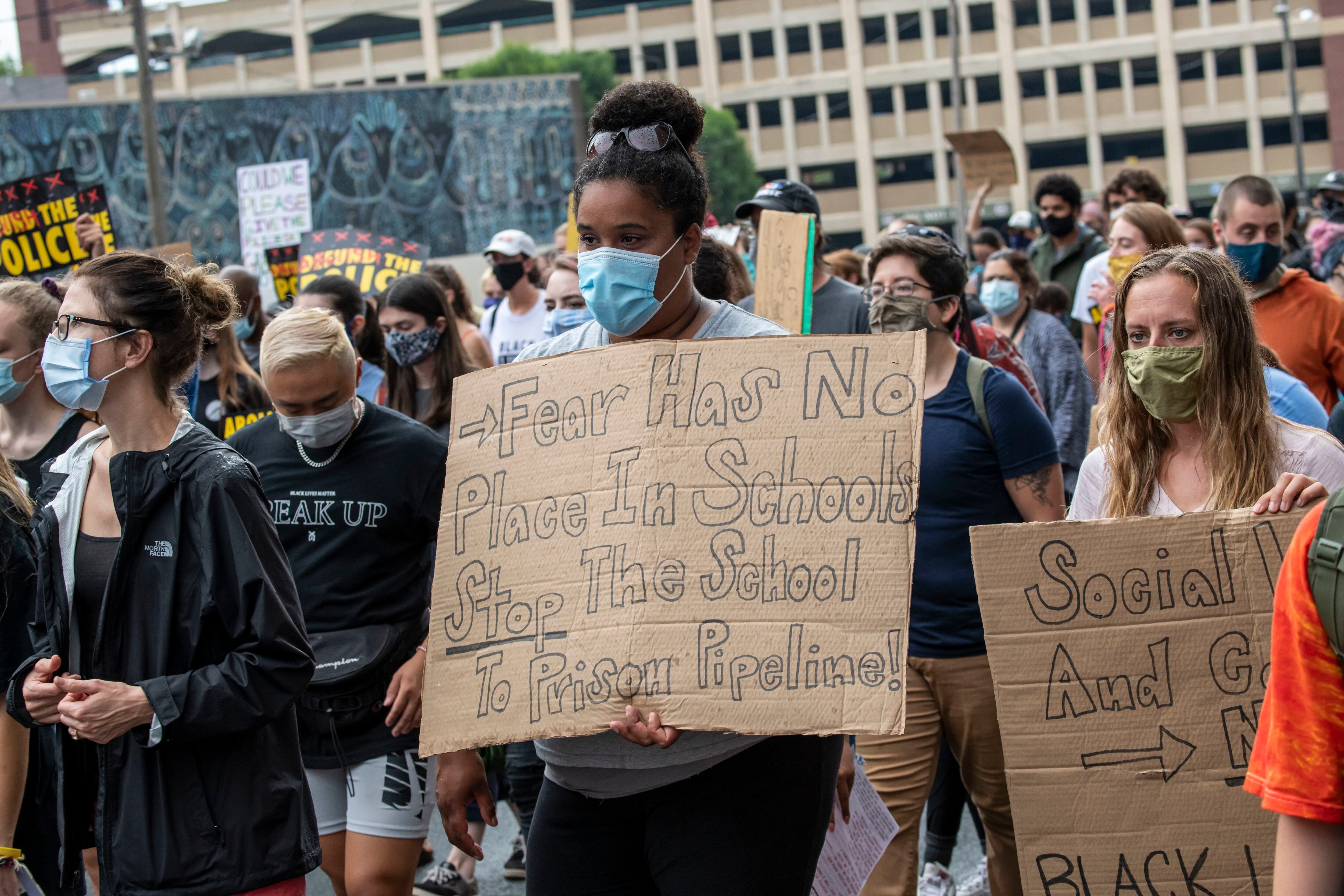 A young woman holds a cardboard sign that reads “Fear has no place in schools, stop the school to prison pipeline!” She is one of many in a large crowd of protestors.