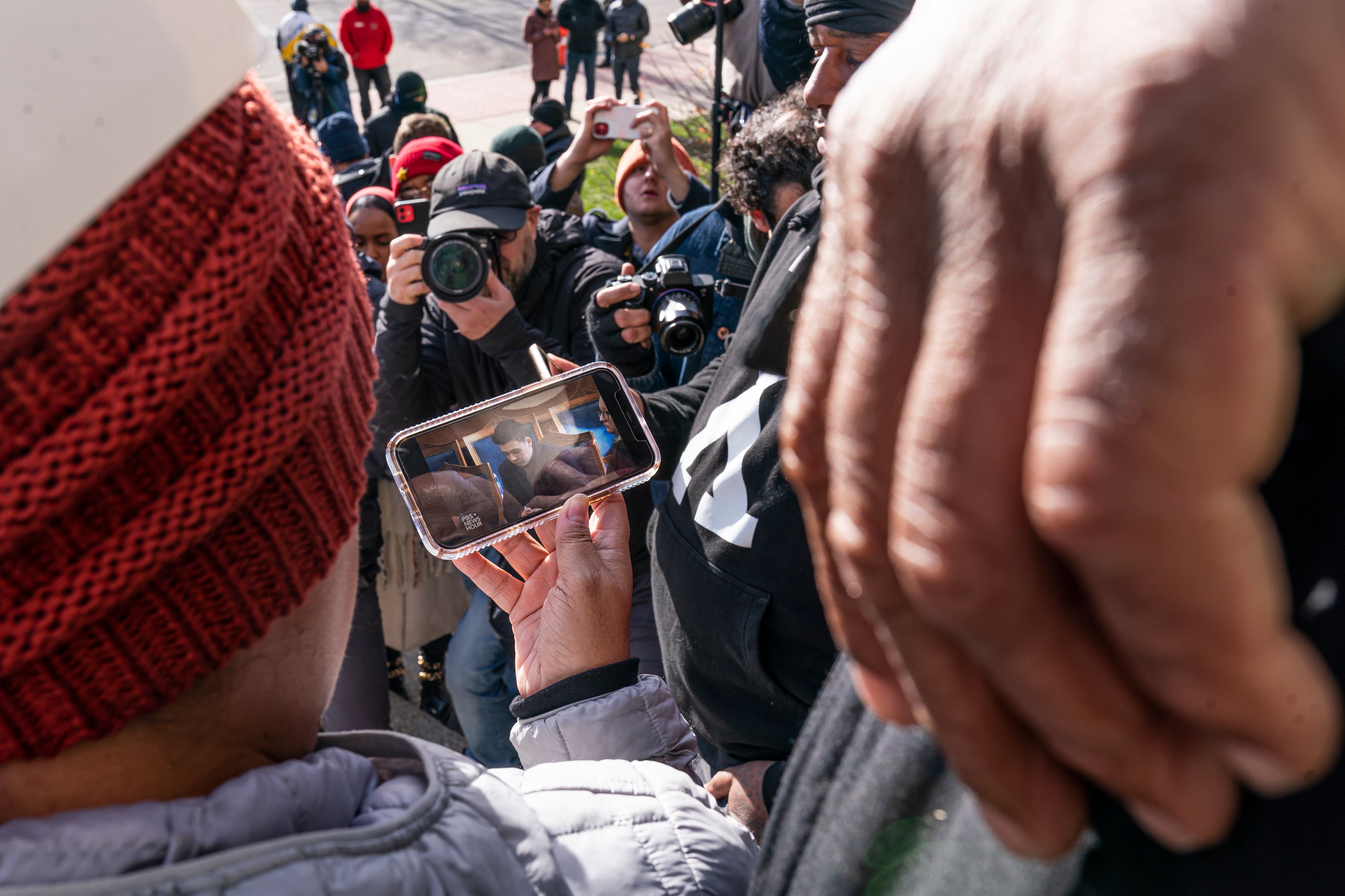 Demonstrators watch the trial of Kyle Rittenhouse on a cell phone outside of the courthouse, as members of the media make photographs of them. A hand takes up the right side of the frame in the foreground of the image.