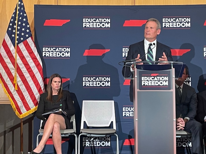 A women sits to the left of a man speaking at a podium with a dark blue background with an American flag to the left.
