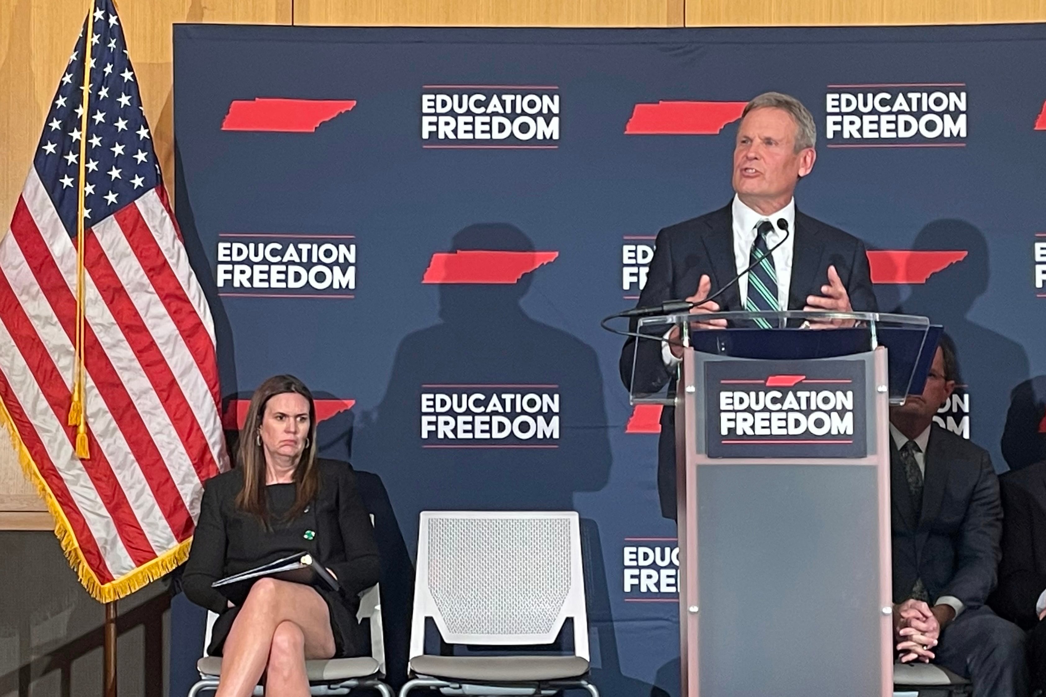A women sits to the left of a man speaking at a podium with a dark blue background with an American flag to the left.