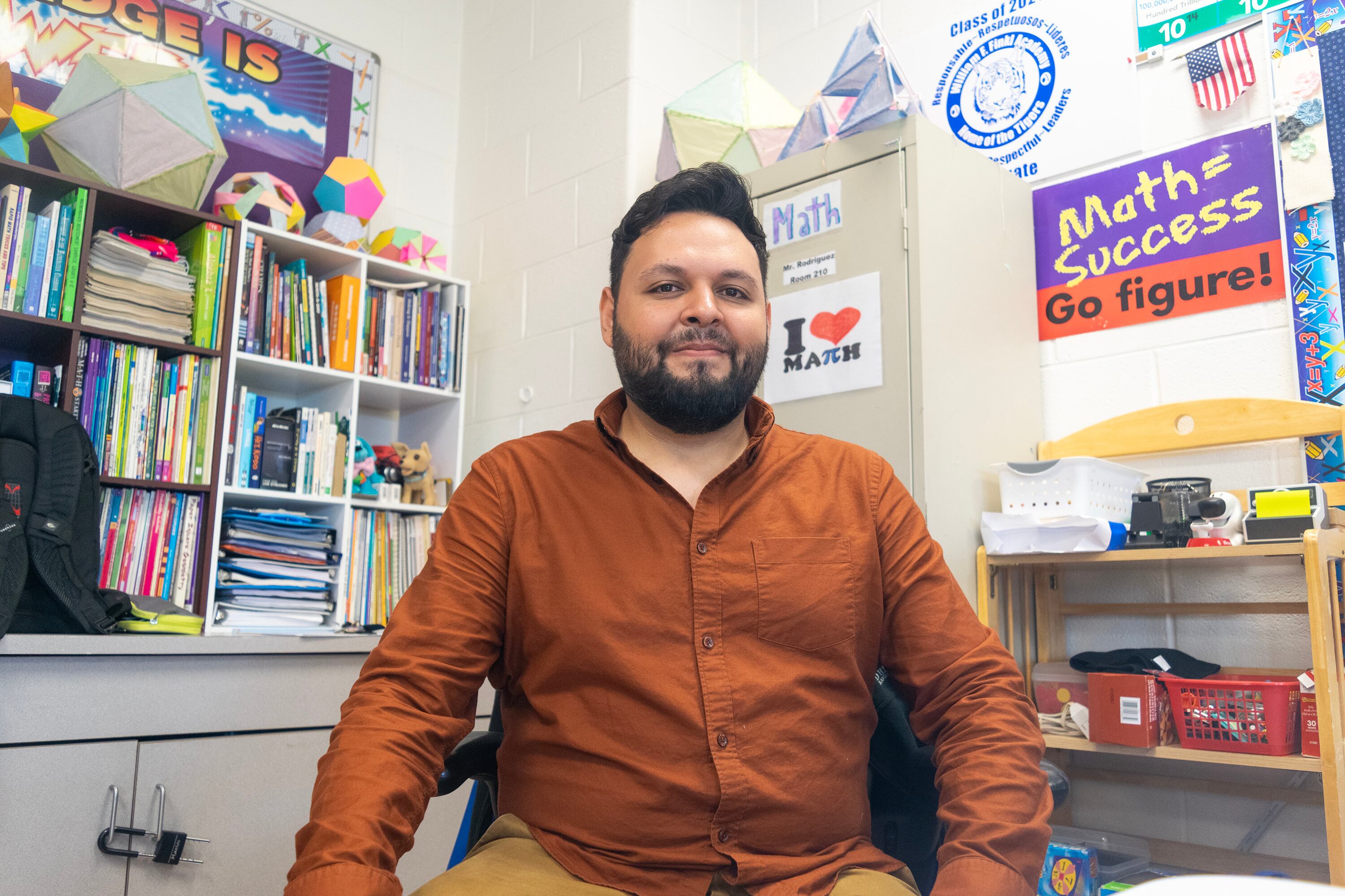 Giovanny Navarro, wearing an orange formal shirt, sits at an office chair. Behind him are signs that say “Math=success” and “I heart math.” There also is a bookshelf and file cabinet behind him with colorful books. Navarro smiles toward the camera