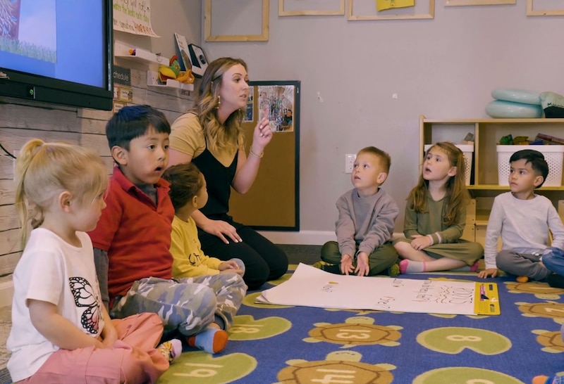 A teacher with blonde hair sits in a circle with young students in an early childhood education classroom.
