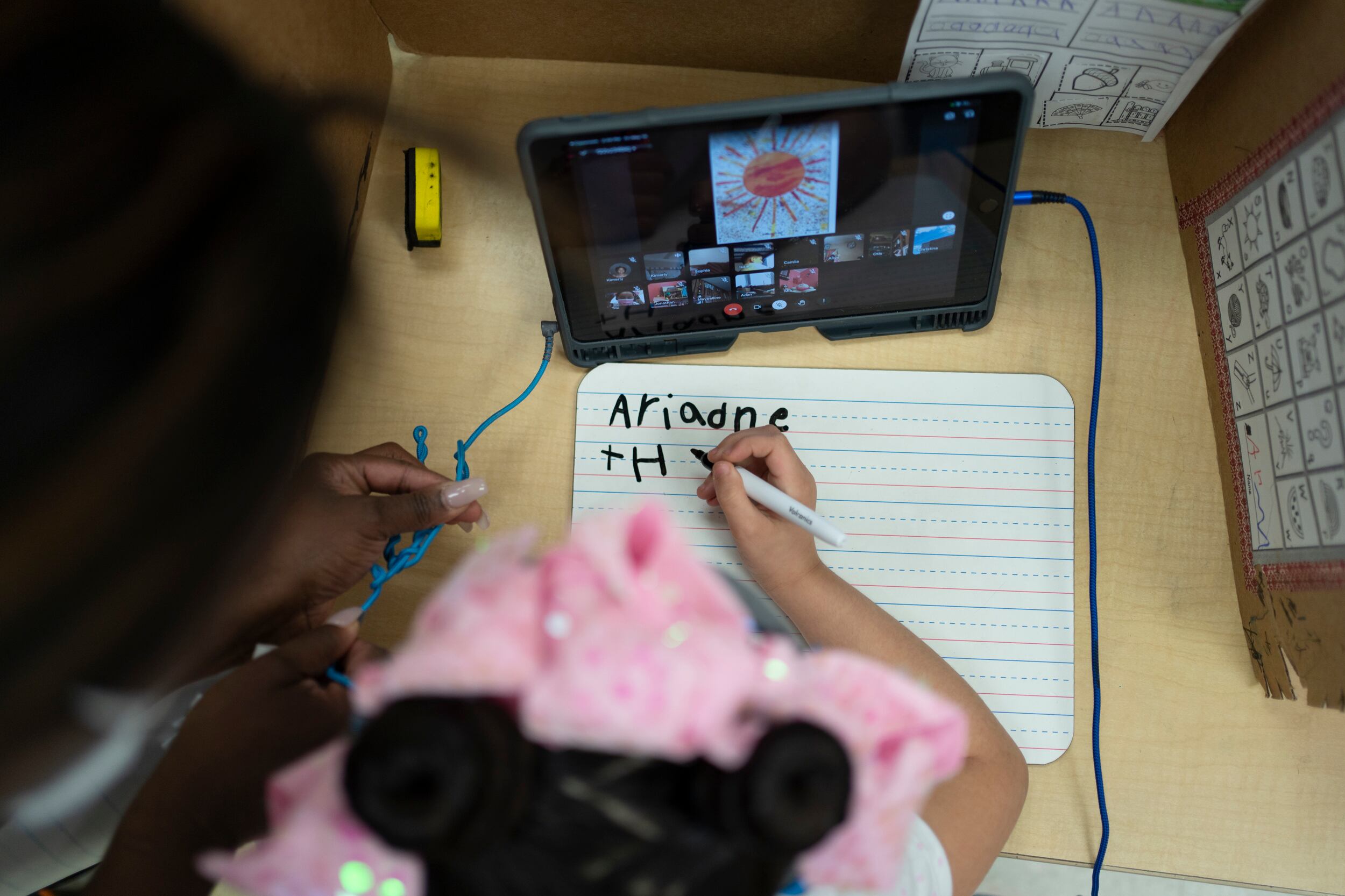 A girl wearing a pink bow works on a writing assignment while a teacher looks over her shoulder. There is a tablet on the desk in front of her.