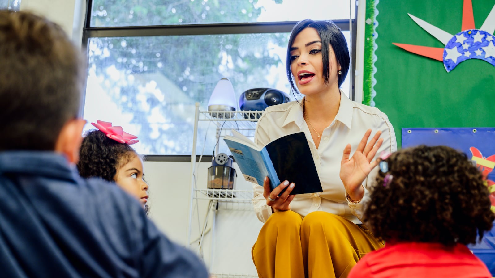 Young female Hispanic schoolteacher reading aloud to young students sitting around her on the floor.