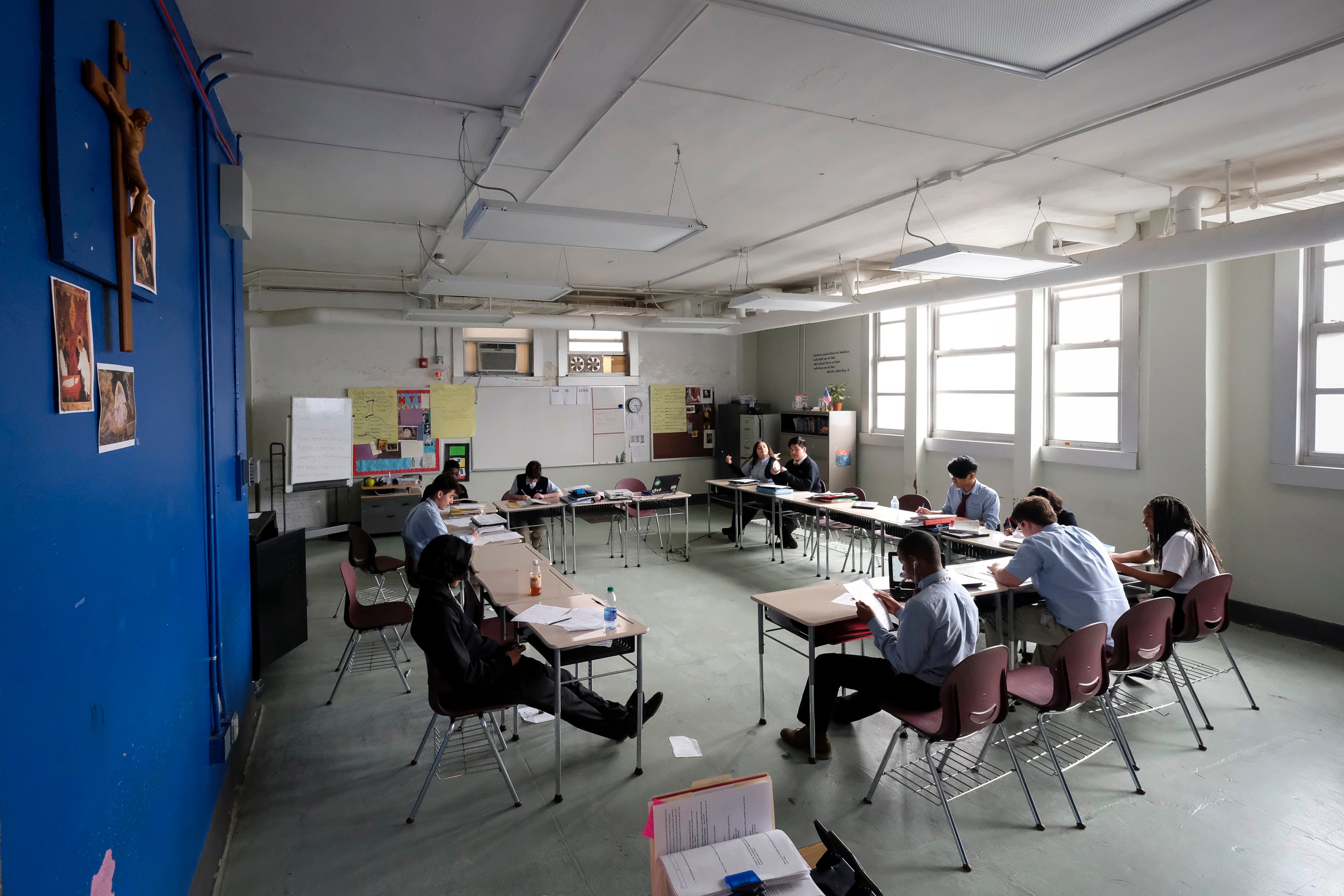 11 high school students look at papers and books while sitting at desks arranged in a large rectangle in an Indiana classroom. Pictures are pinned on a royal blue wall, and more pictures and posters are pinned to bulletin board on a far wall.