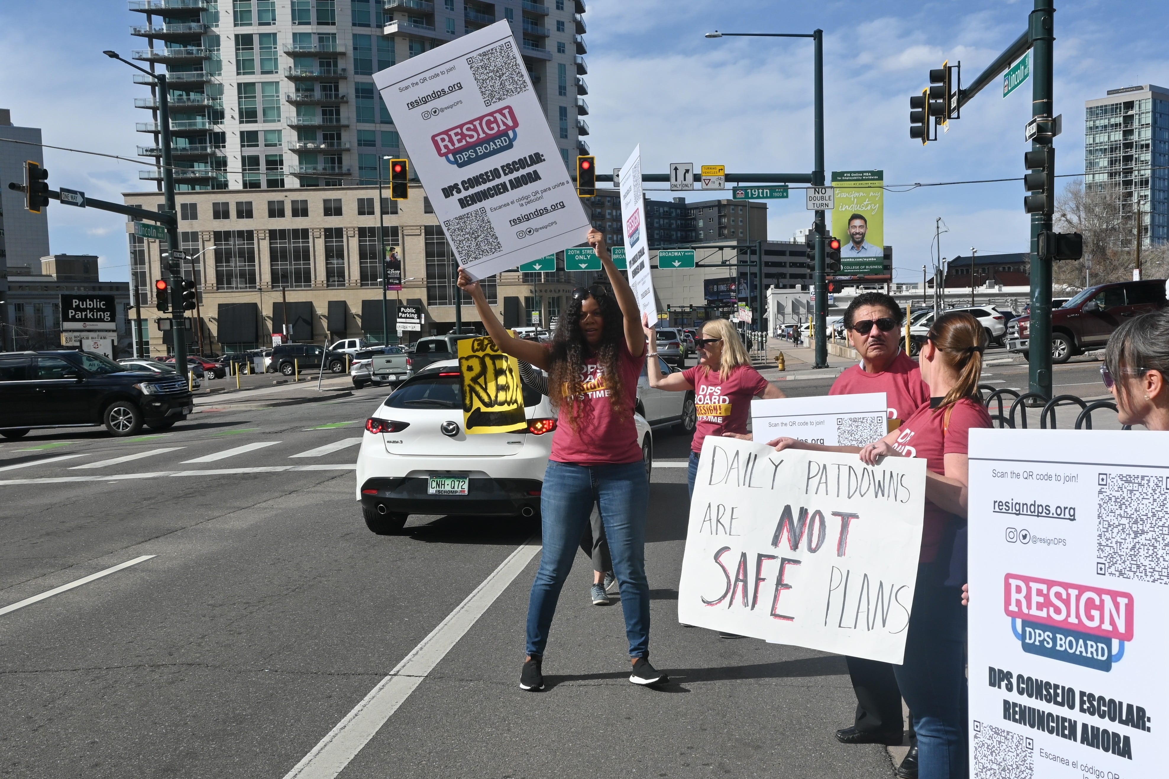 A woman dressed in a red shirt holds up a sign calling for the Denver Public Schools board to resign while cars pass by.
