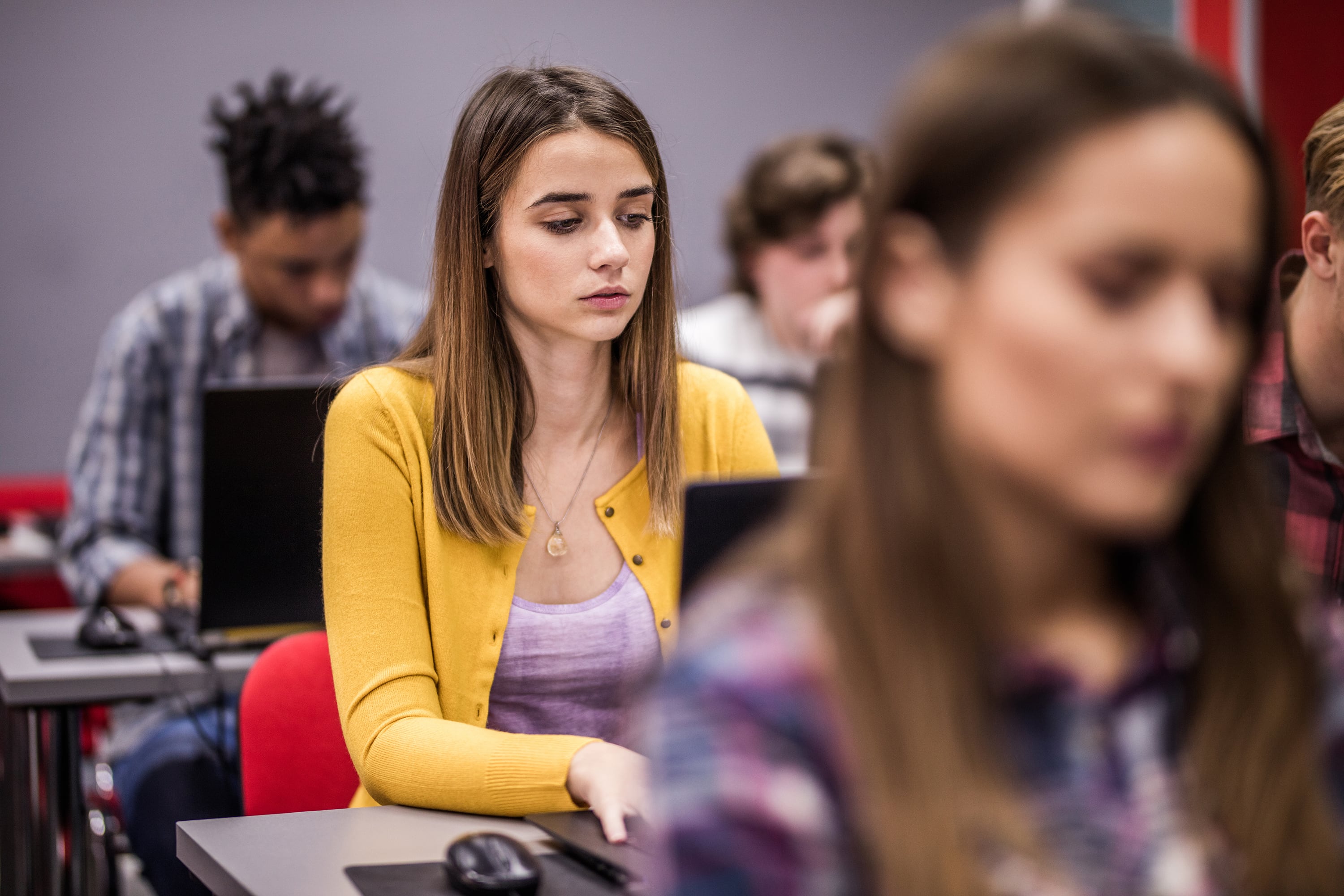 One student with long brown hair and wearing a bright yellow sweater sits at a desk working on a laptop is in focus between two rows of students in the foreground and background.