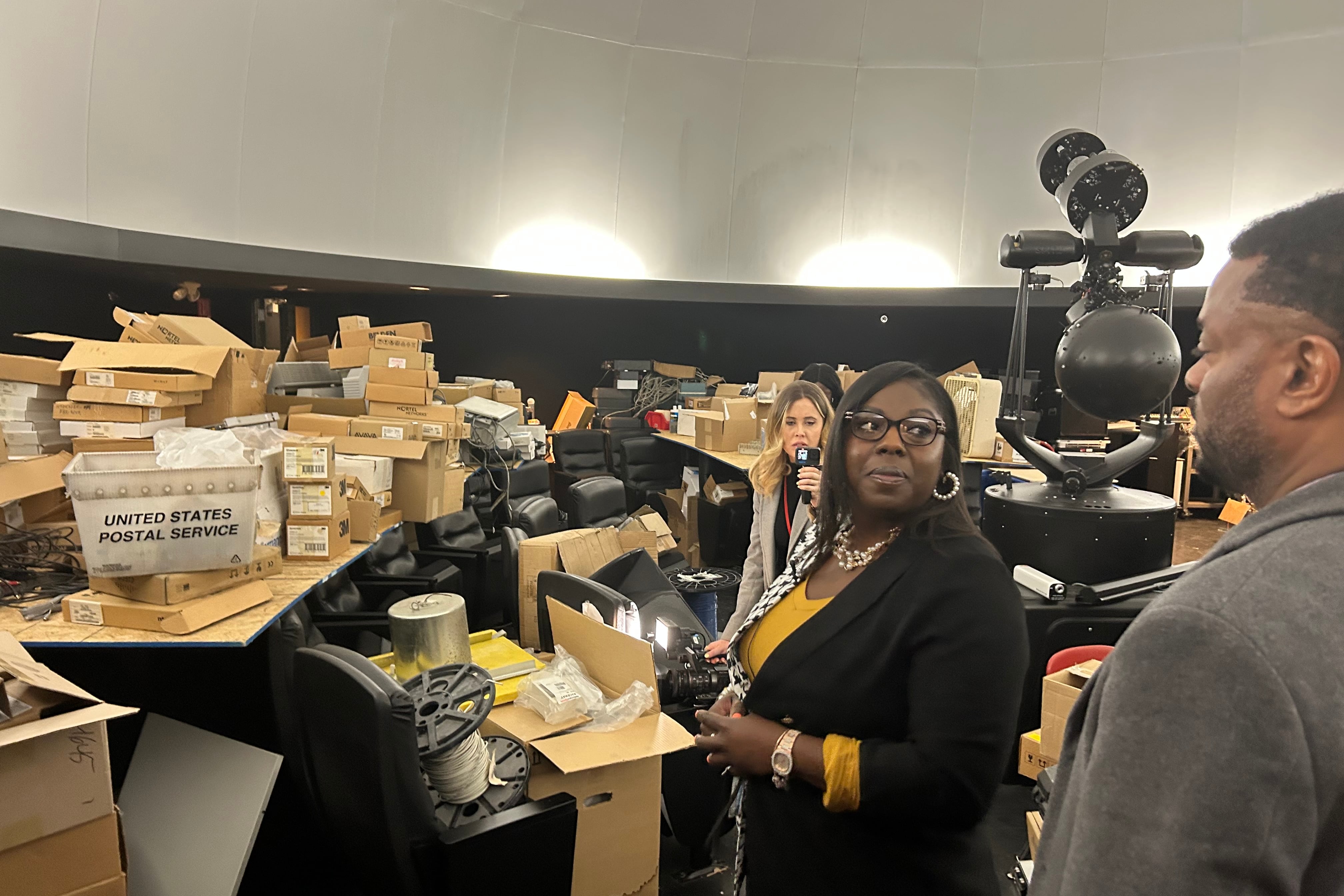 A woman wearing glasses, a yellow shirt, and black jacket stands in front of a juble of chairs, boxes, and other equipment.