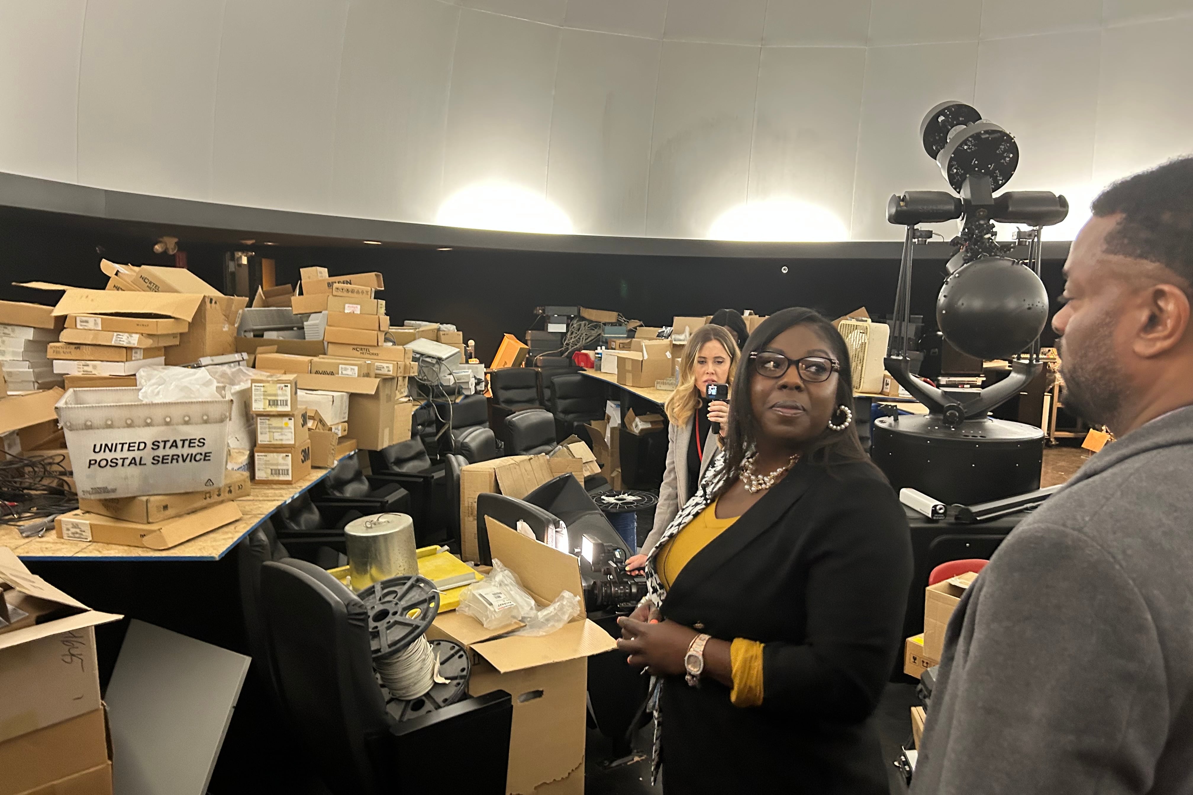 A woman wearing glasses, a yellow shirt, and black jacket stands in front of a juble of chairs, boxes, and other equipment.