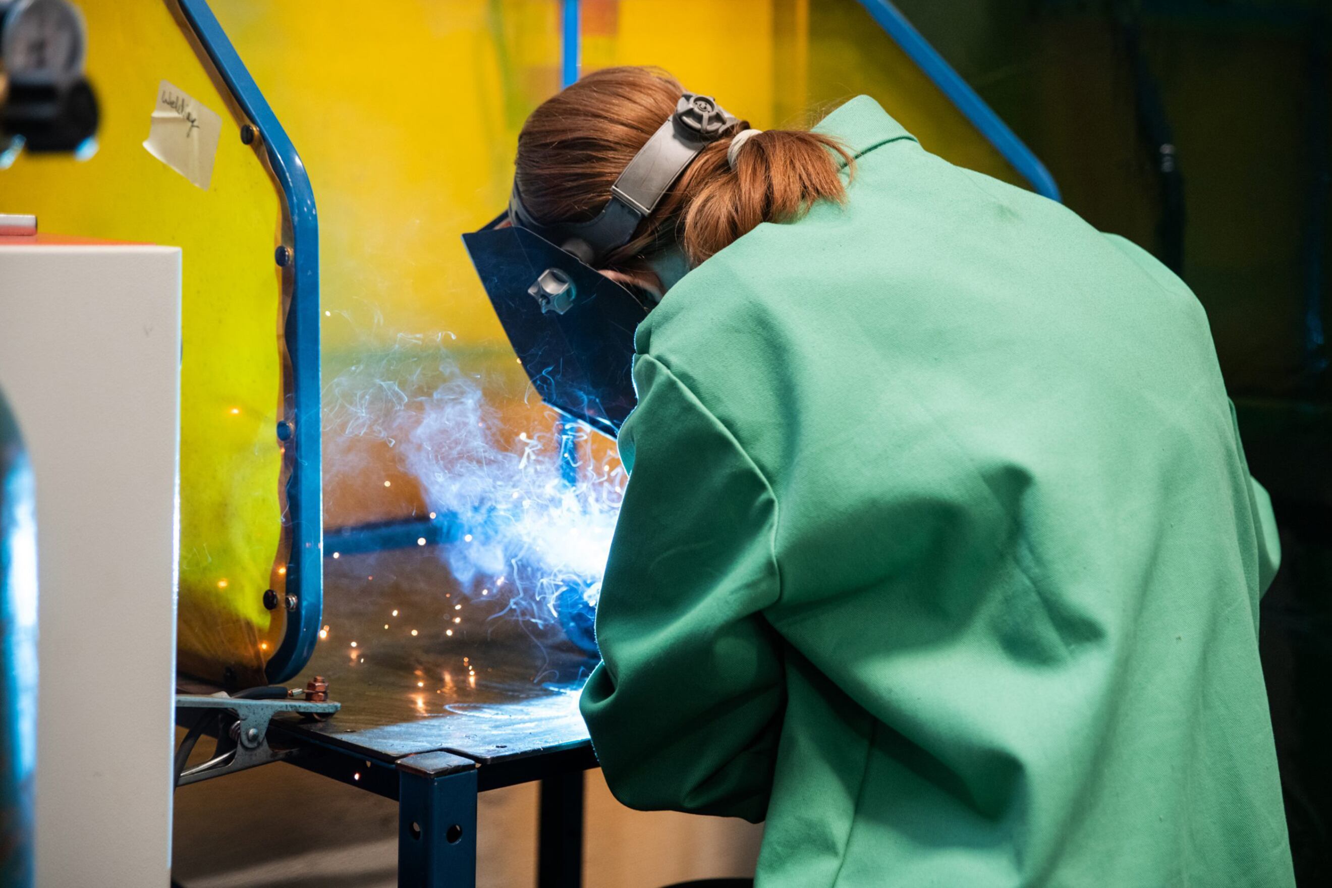 A young woman with red hair, goggles and green smock welds.