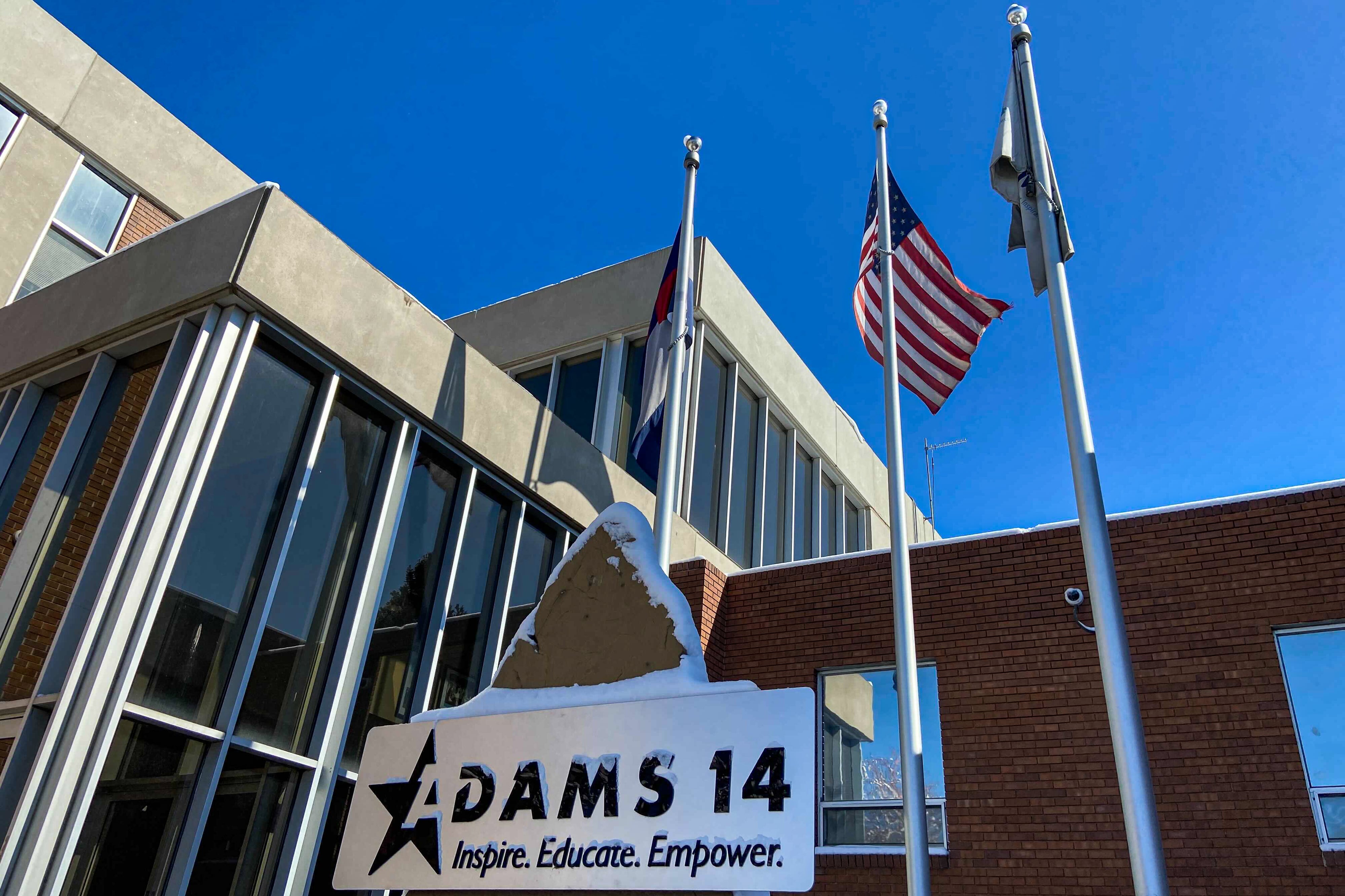 Flags fly outside the Adams 14 school district headquarters. The district logo sits prominently in front of the building. The sky is a clear blue.