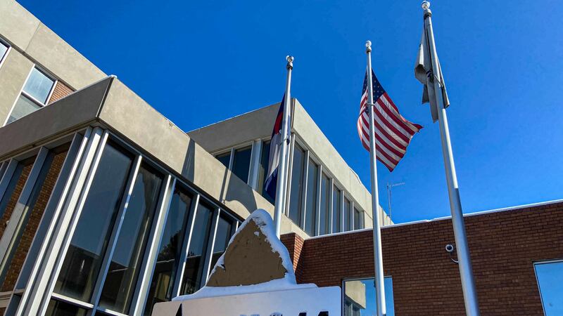 Flags fly outside the Adams 14 school district headquarters. The district logo sits prominently in front of the building. The sky is a clear blue.
