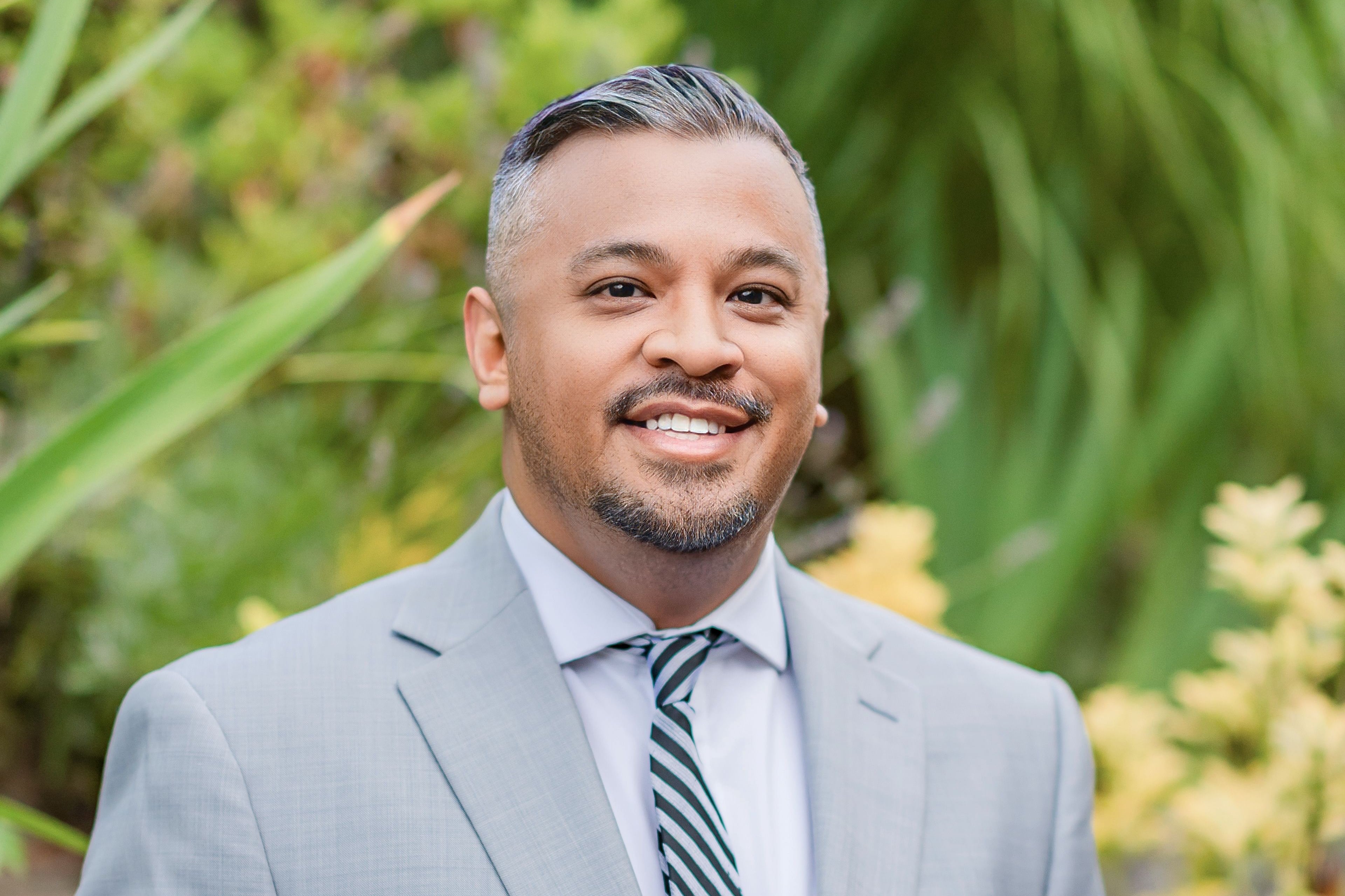 A man smiles in a light gray suit and black-and-gray tie.