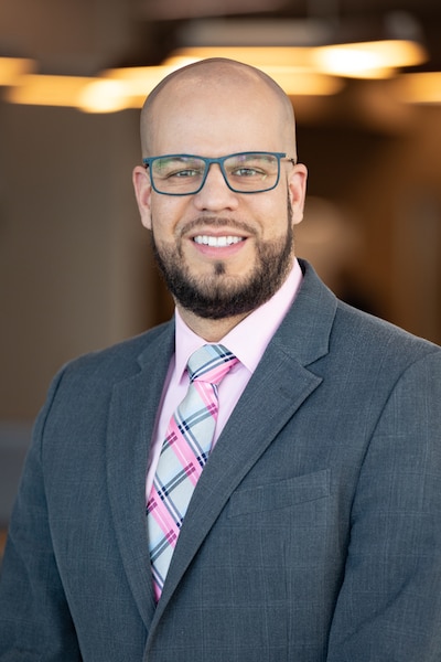 Headshot of a man, wearing a gray suit, a multicolored tie, and glasses.