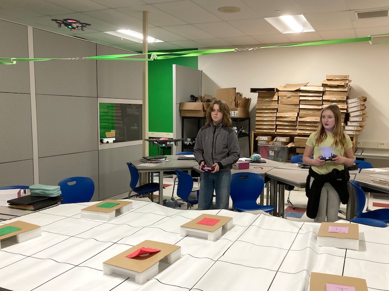 Two middle students stand next to each other in a classroom flying drones.