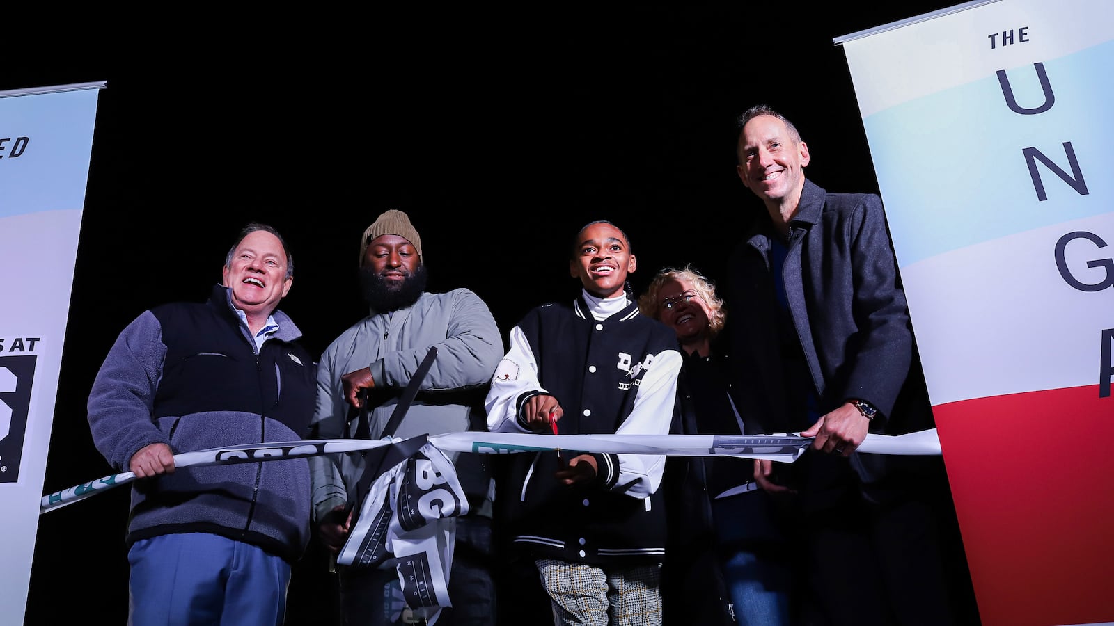 A young man and four adults perform a ribbon cutting during a facility unveiling, bright lights shining on them on a dark night.