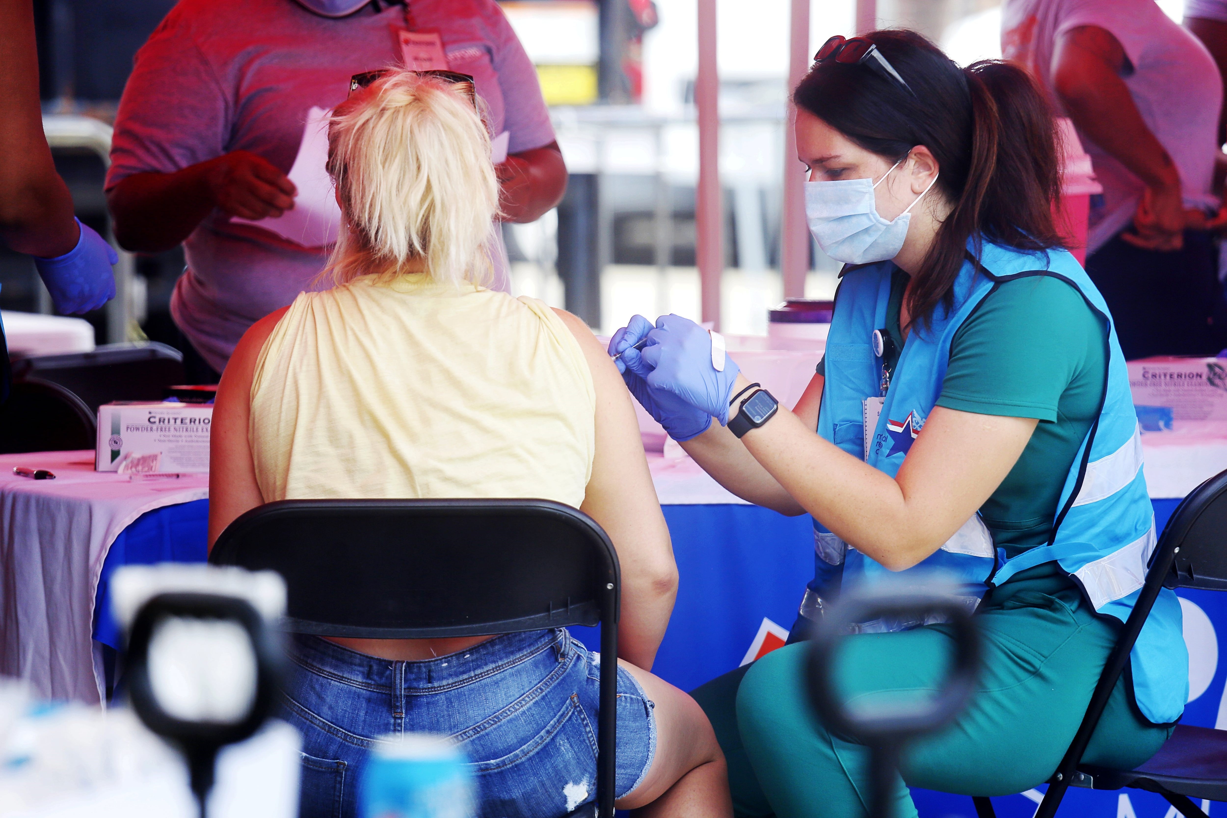 A woman wearing a yellow shirt seated on a black metal folding chair receives a shot in the arm from a nurse wearing a face mask and gloves.