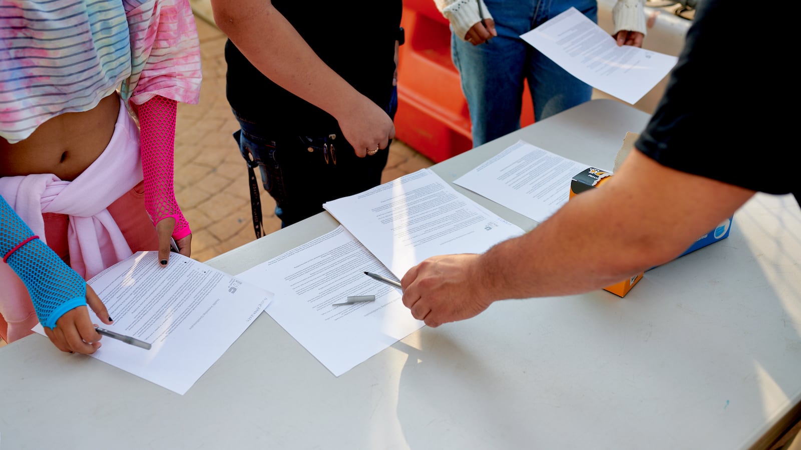 Students receive COVID screening forms and pens at a table from faculty.