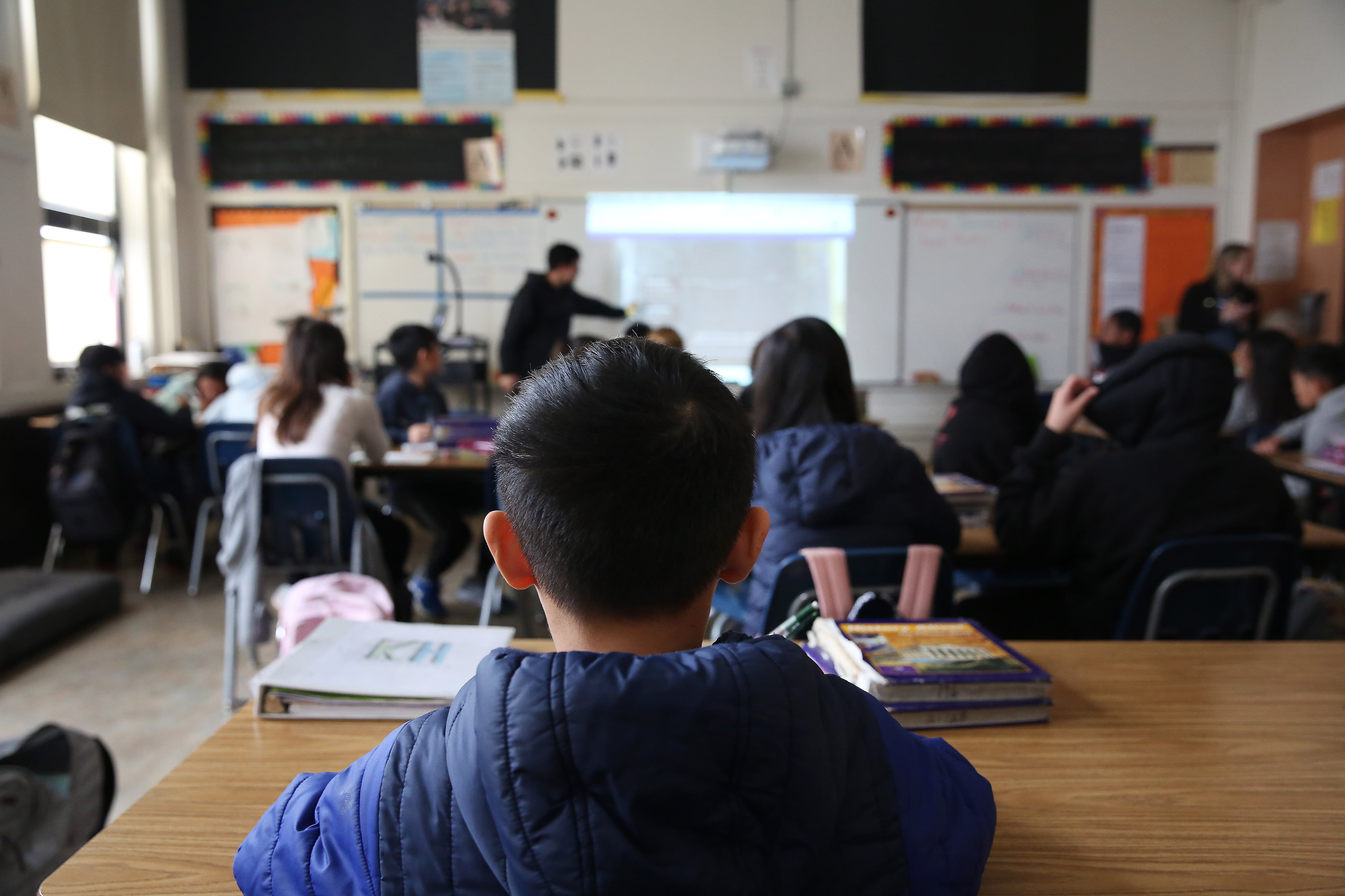 A young boy with short hair sits at a school desk looking ahead and watching as a teacher points to a whiteboard.