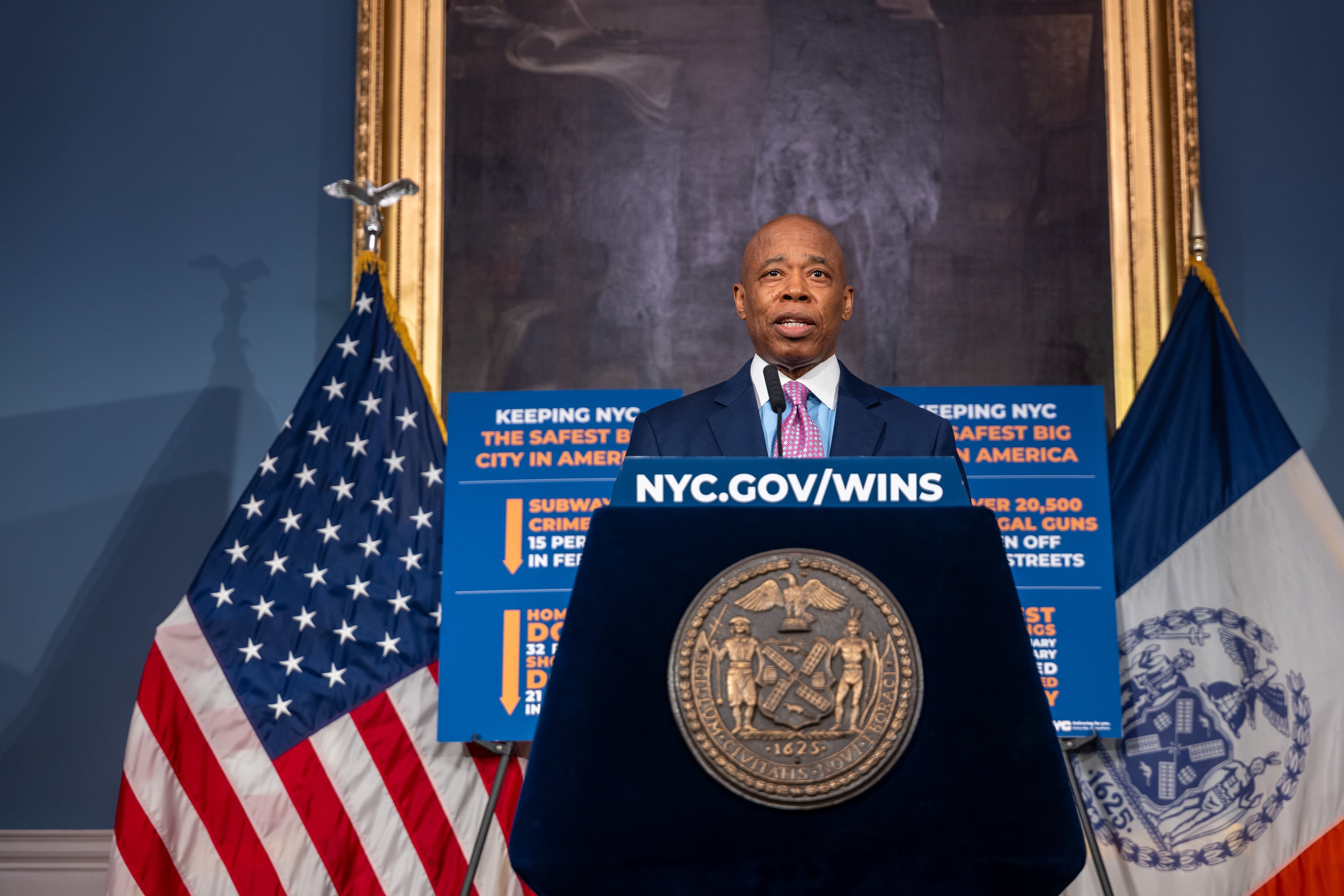 New York City Mayor Eric Adams speaks from behind a podium and in front of a wall with a large painting and two flags in the back.