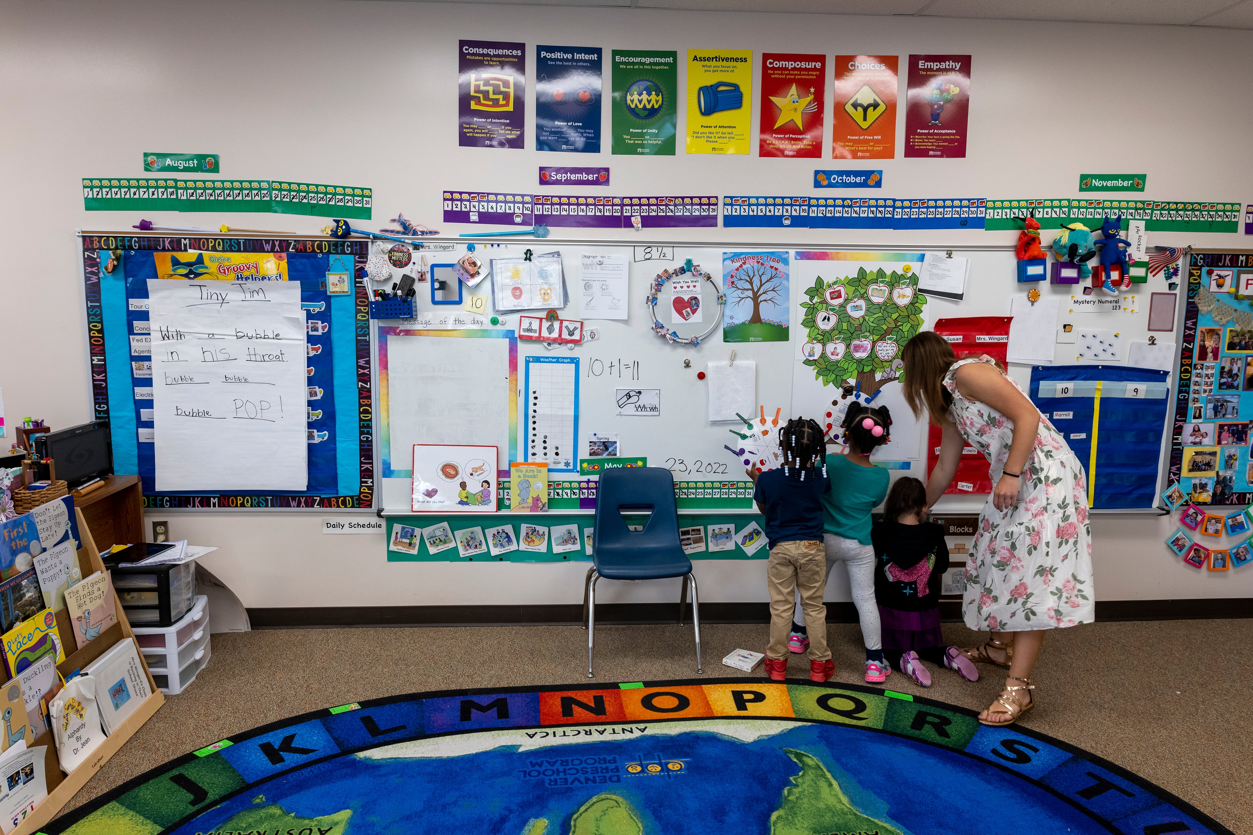 A woman in a flowered dress looks at a classroom bulletin board with three small children.