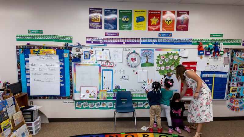 A woman in a flowered dress looks at a classroom bulletin board with three small children.