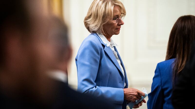 Betsy DeVos wearing a blue blazer looks slightly down as she stands in a listening posture