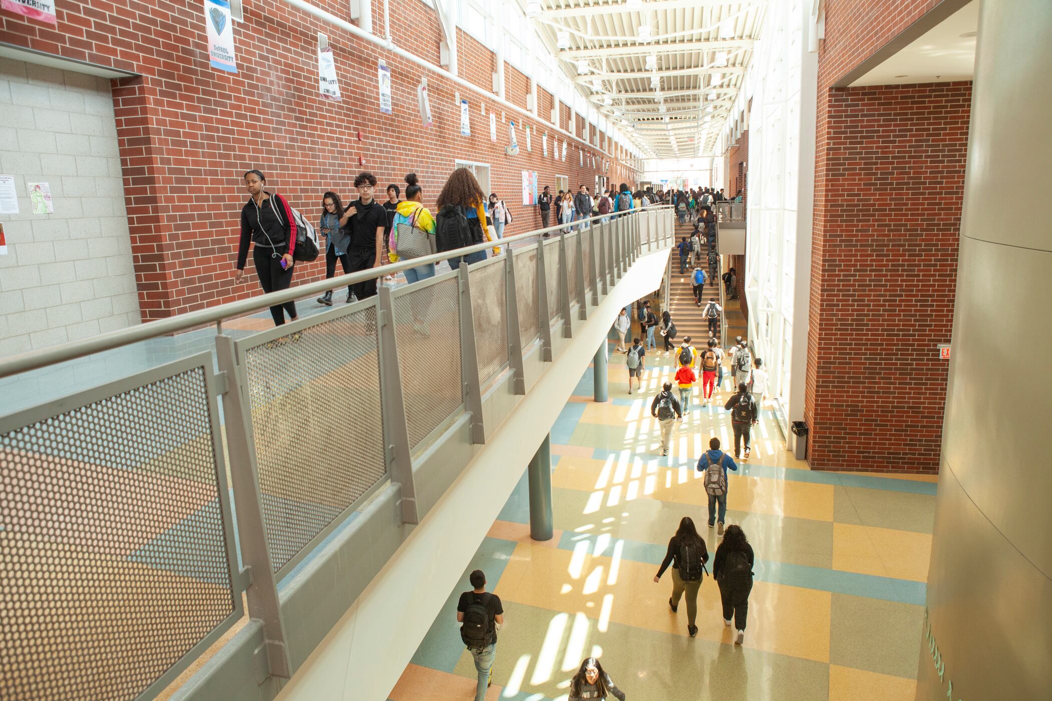 Students walk on a second floor walkway and down a hallway on the ground level in a large high school with lots of natural light coming through many windows.