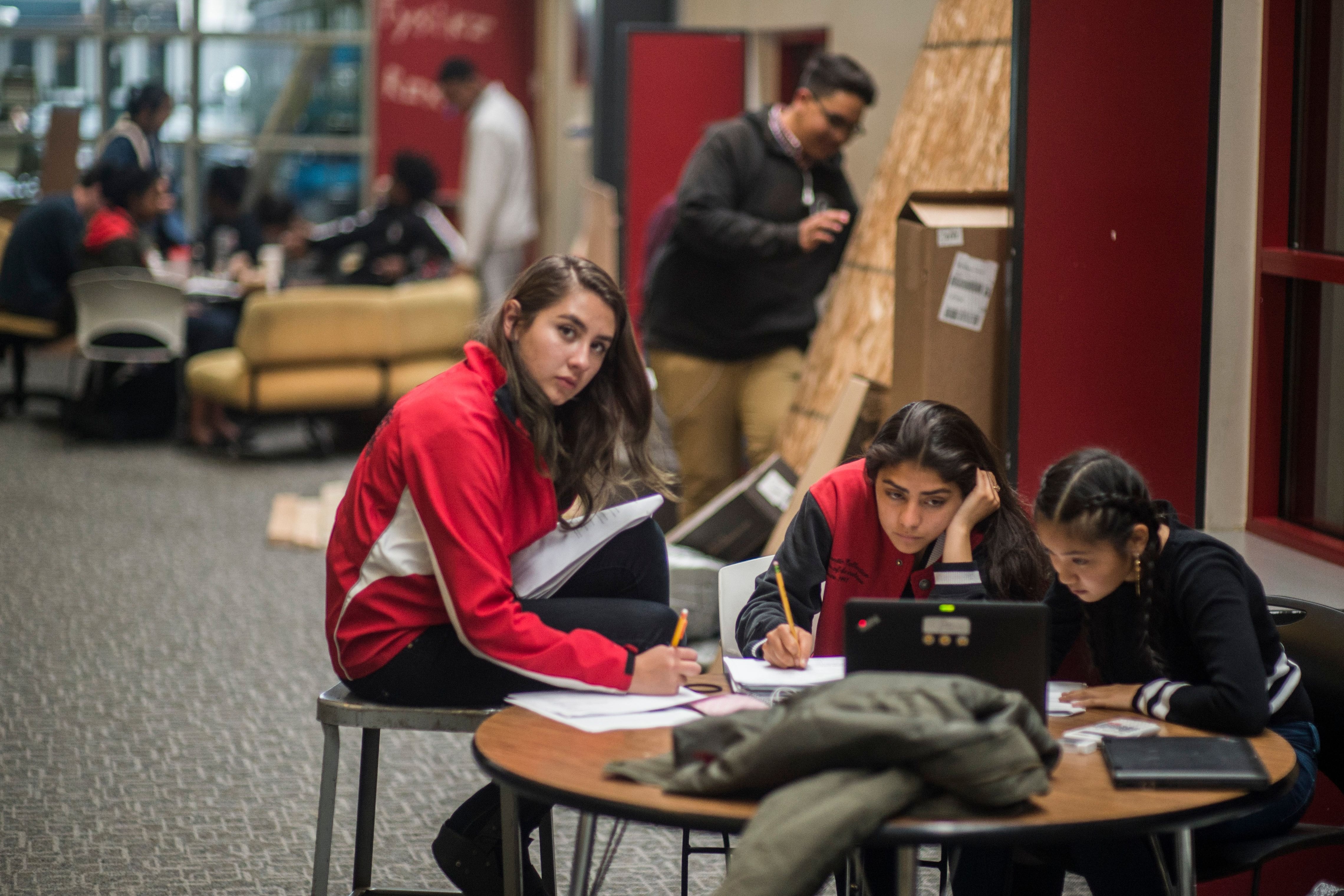 A girl in a red jacket and two other students sit at a round table writing on papers and looking at a laptop. A male stands in the background, and a half dozen others sit and stand in a corner of chairs in a wide hallway.