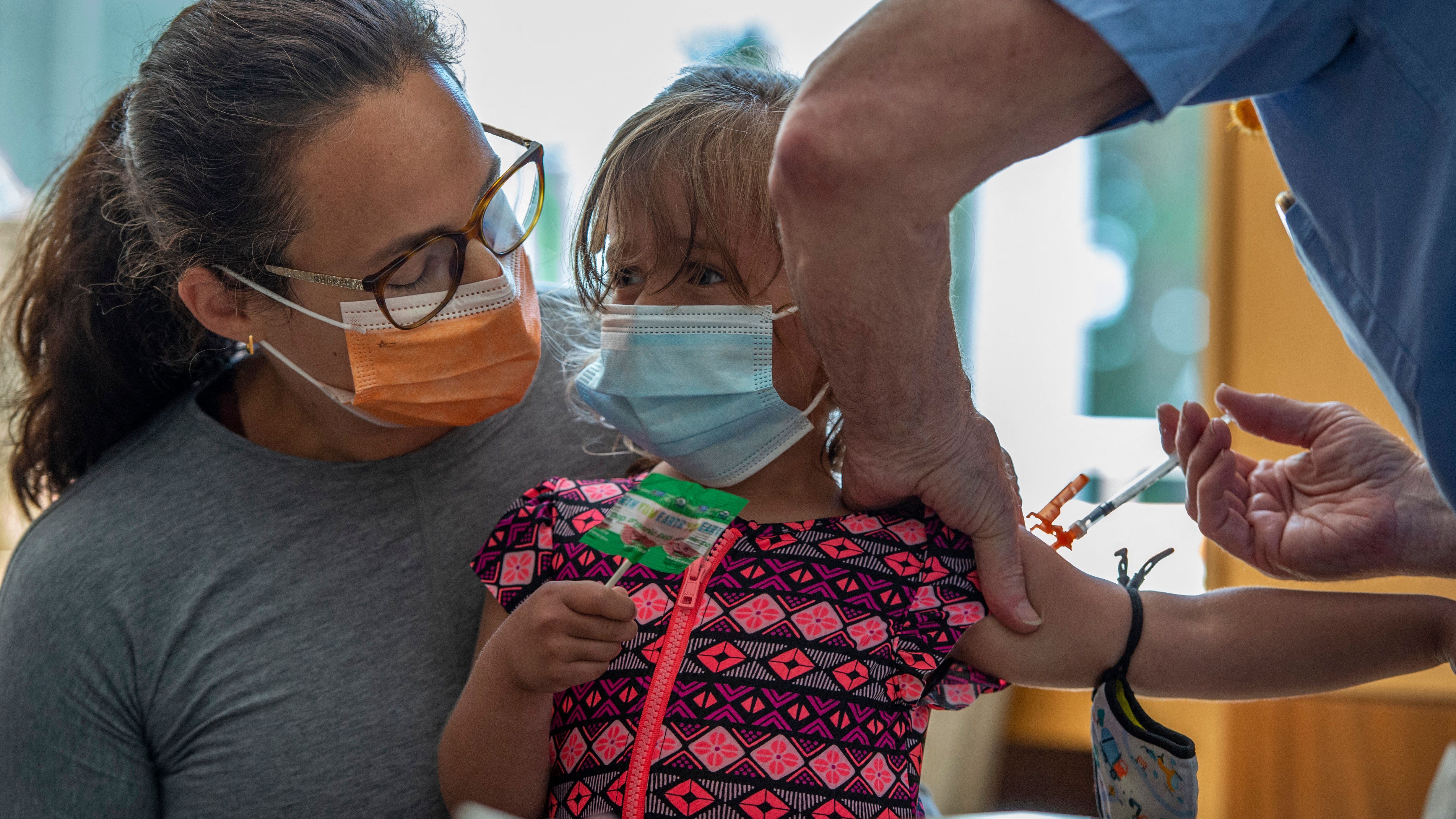 [From left to right] A woman in a gray shirt and orange mask holds a child in a pink and blue shirt who is receiving a shot. The child is holding a lollipop while a medical professional in blue gloves holds her right arm.