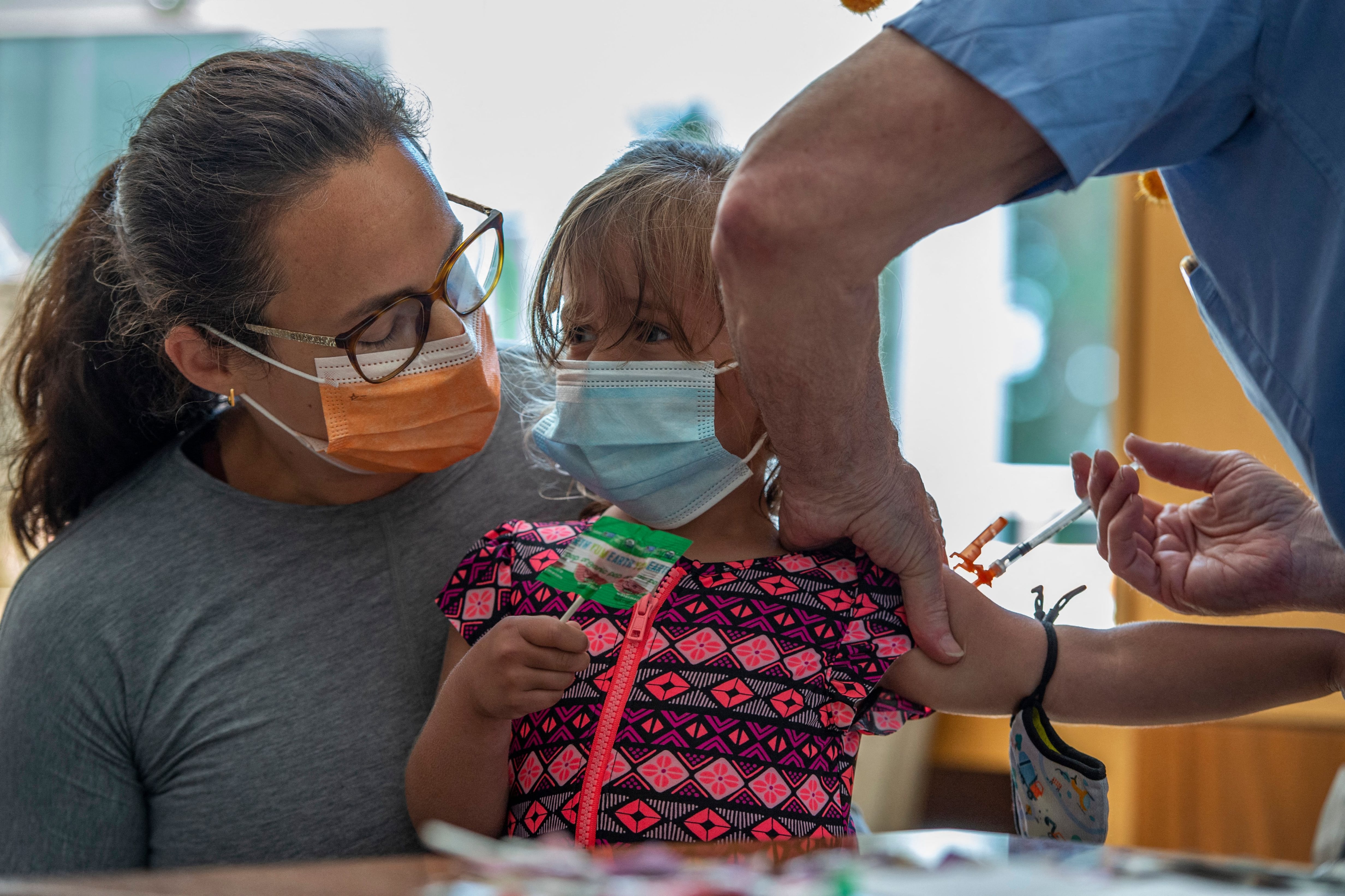 [From left to right] A woman in a gray shirt and orange mask holds a child in a pink and blue shirt who is receiving a shot. The child is holding a lollipop while a medical professional in blue gloves holds her right arm.