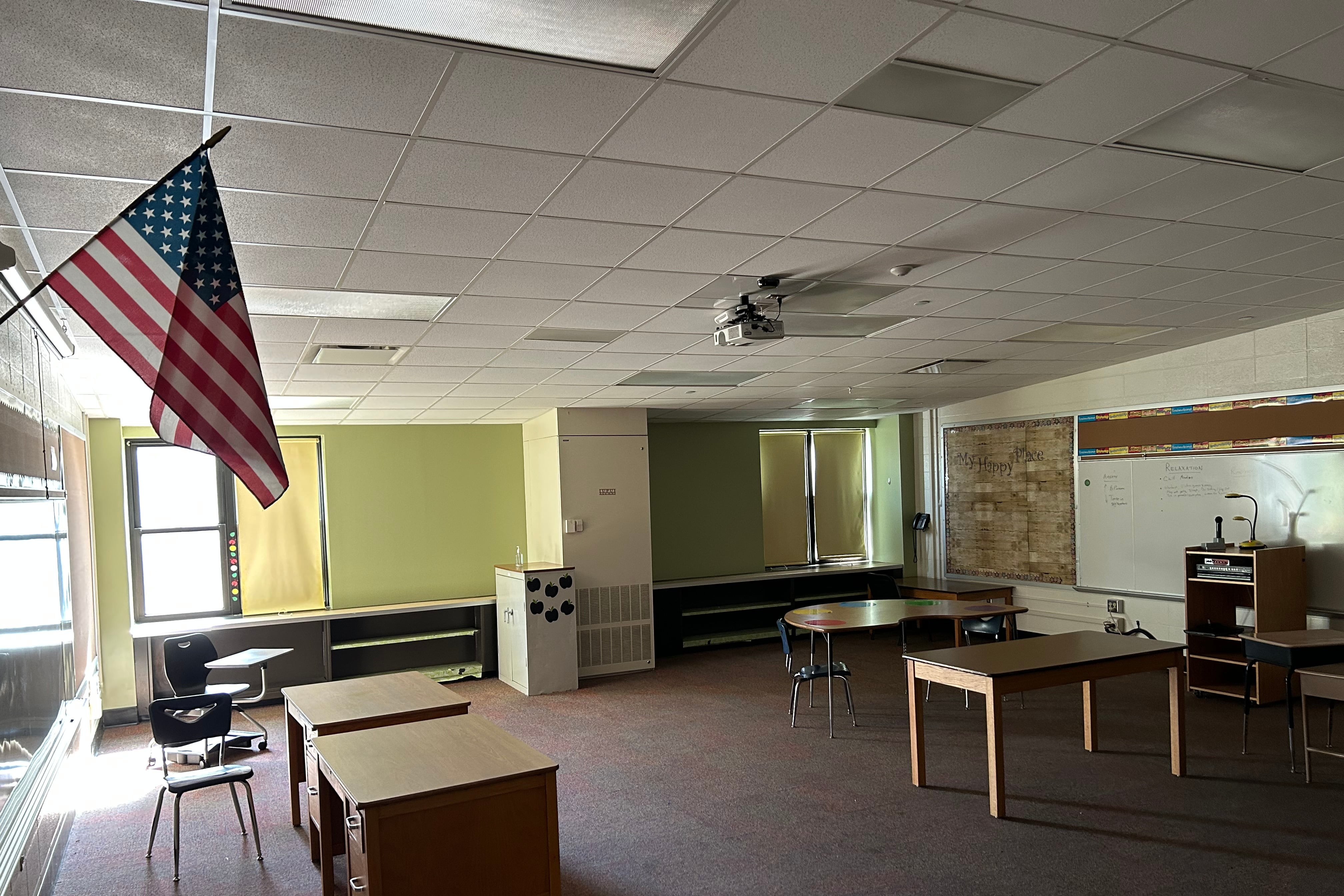 Sun peeks through a window into an empty classroom with an American flag hanging on the wall in the foreground.