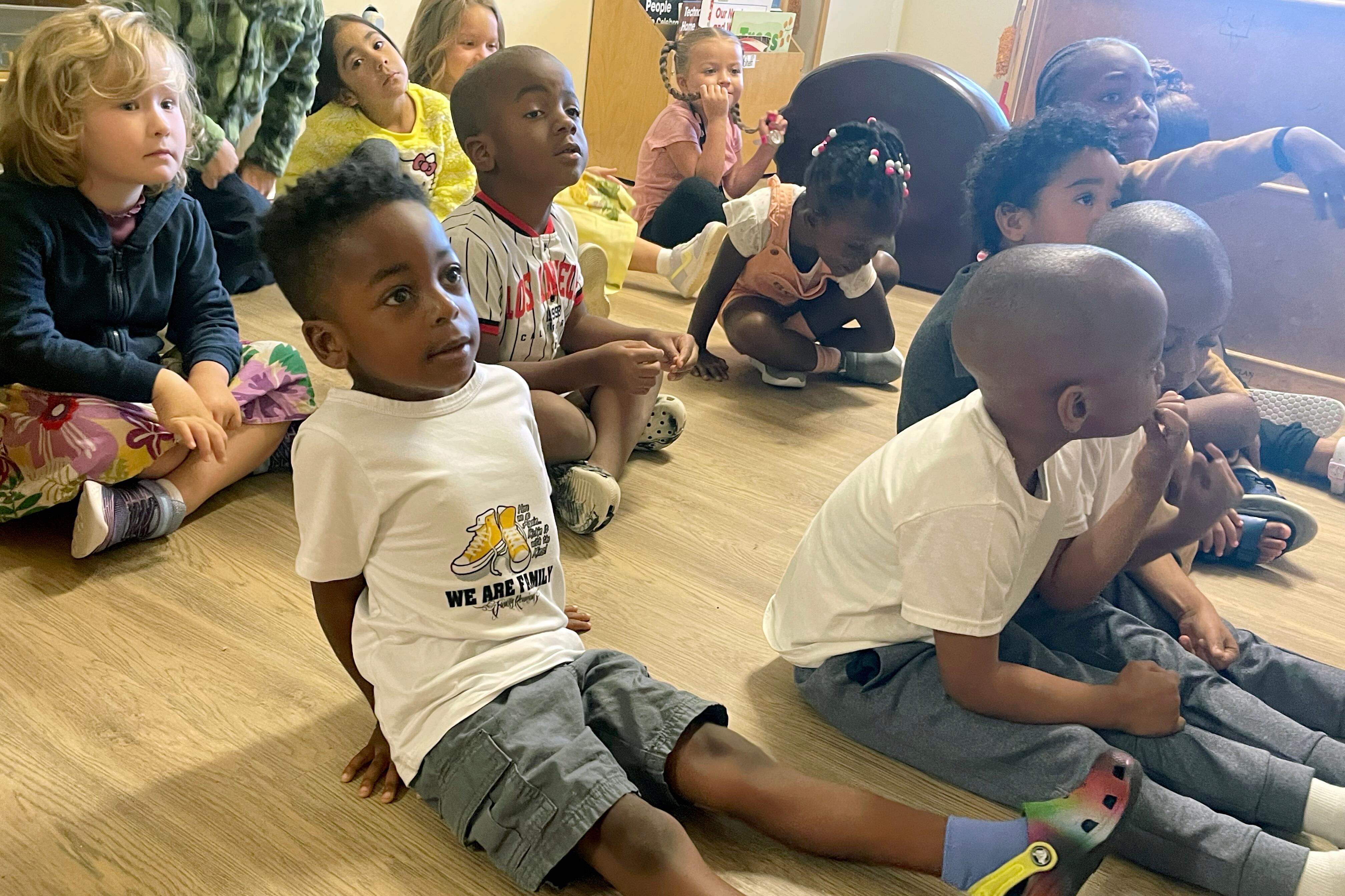 A class of young students sit on the wooden floor in a classroom.