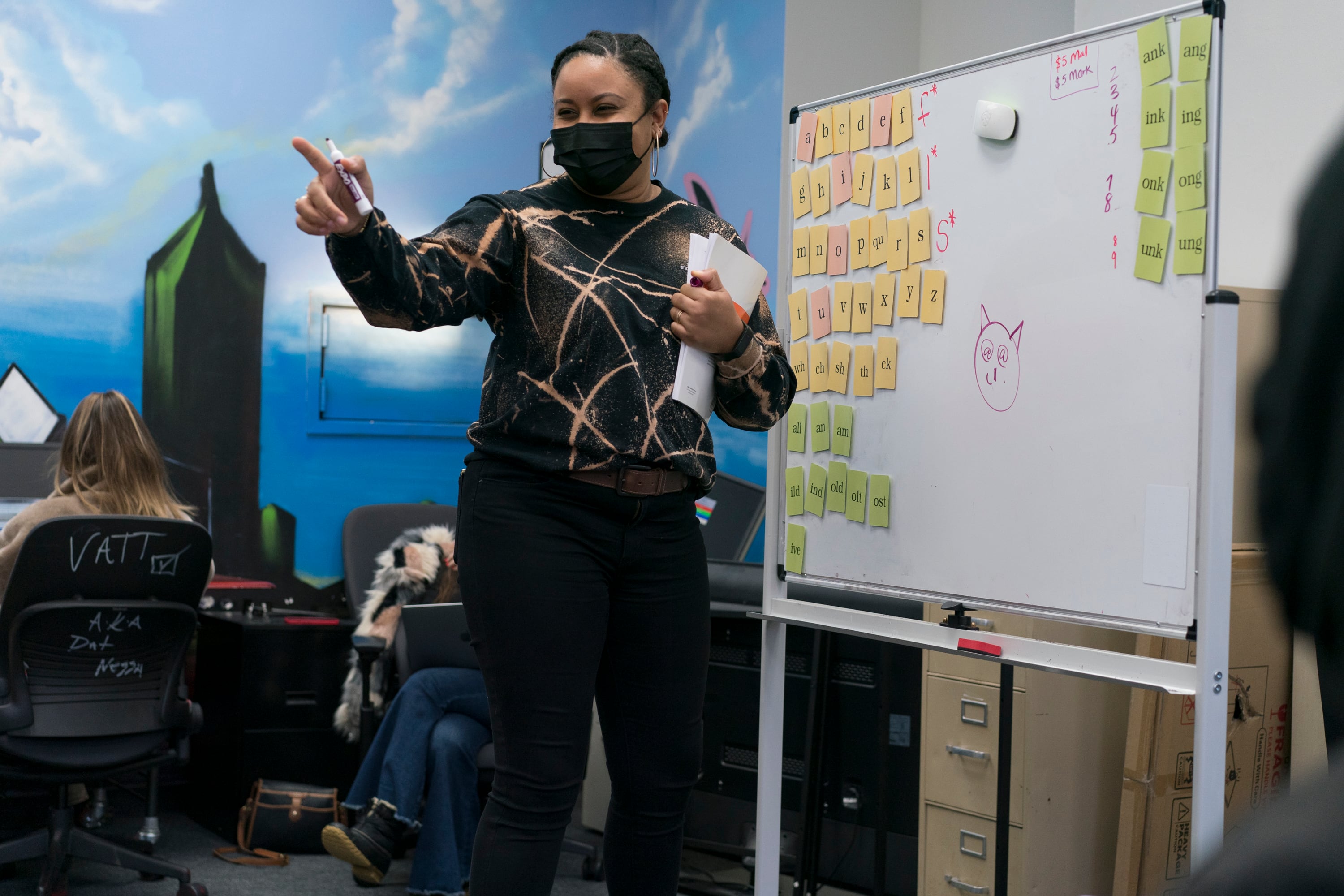 A teacher, wearing a black mask with a black and tan sweater, points elatedly toward a student during reading instruction.