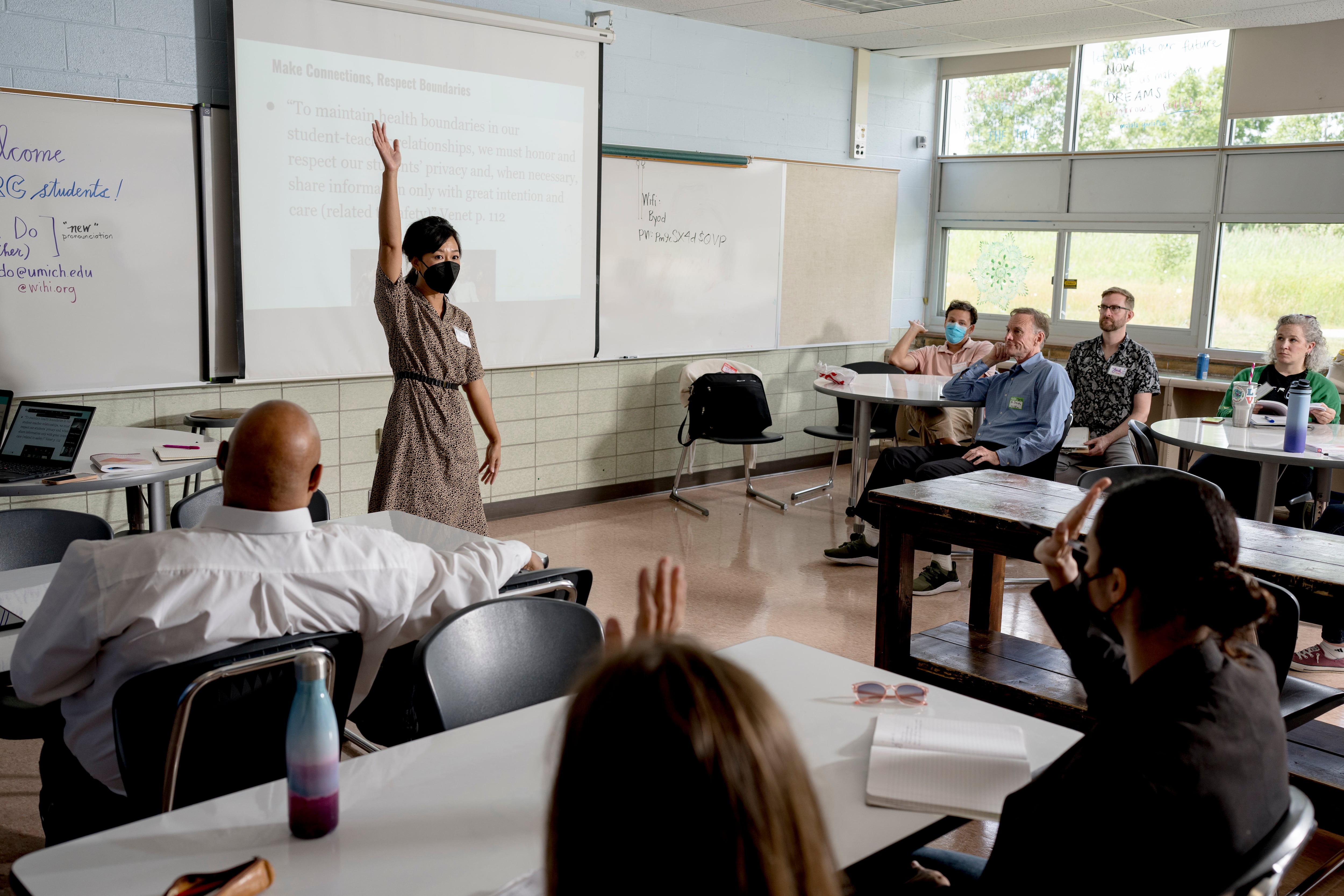 A woman in a dress and wearing a mask raises her hand in front of a project screen while several people sit at desks and look at her.