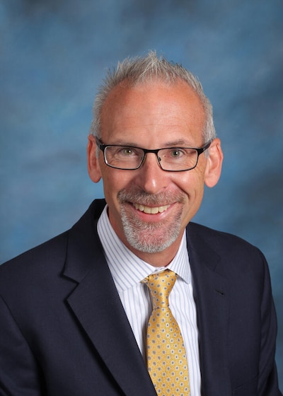 A man wearing glasses and a dark suit with a yellow tie poses for a portrait in front of a light blue and white background.