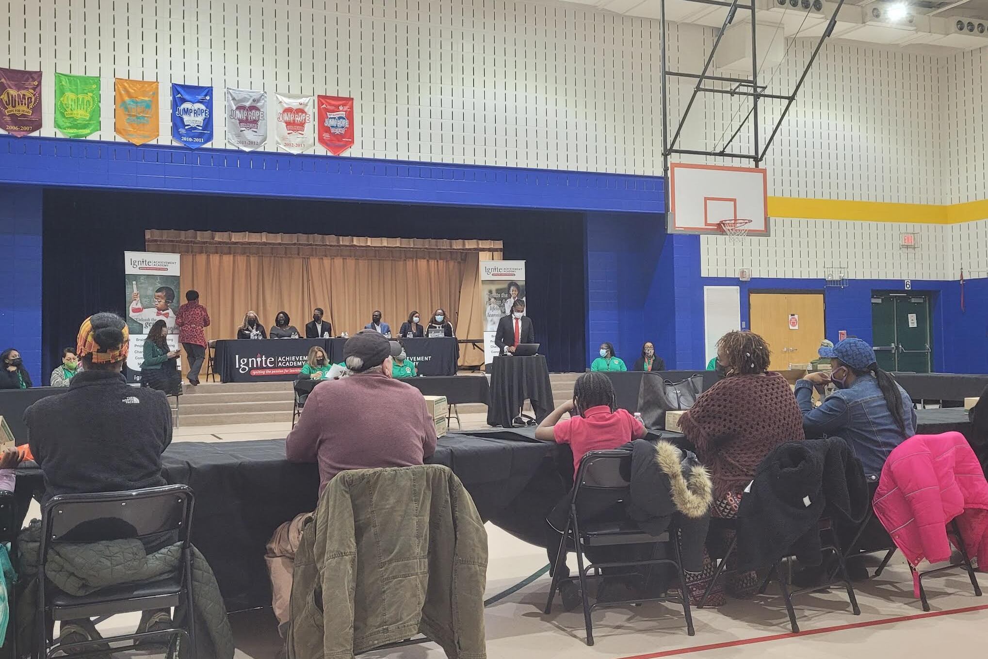 Four parents with their backs turned sit in a gym facing Ignite Achievement Academy co-founder Shy-Quon Ely as he gives a speech on the status of the school, with board members on a raised stage behind him.