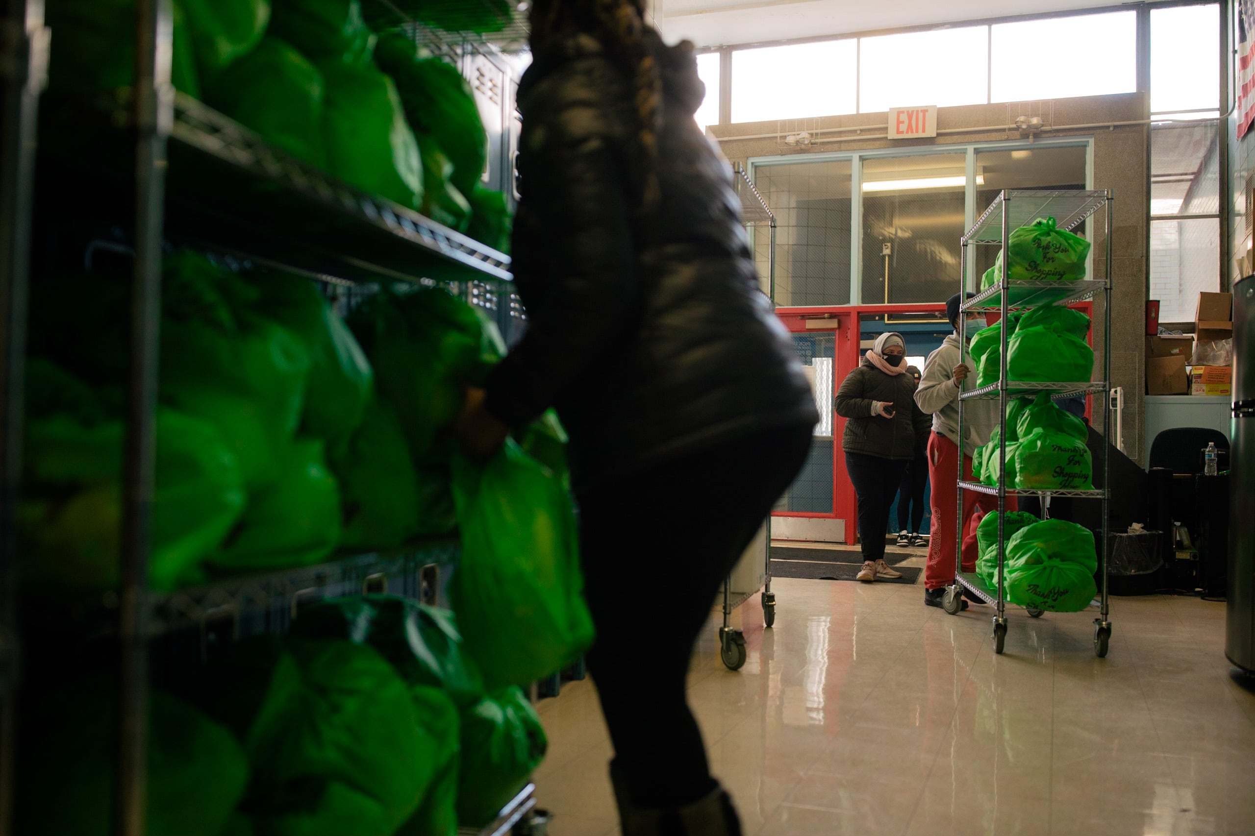 Three adults work in a school food pantry next to large wire racks with green bags full of food.